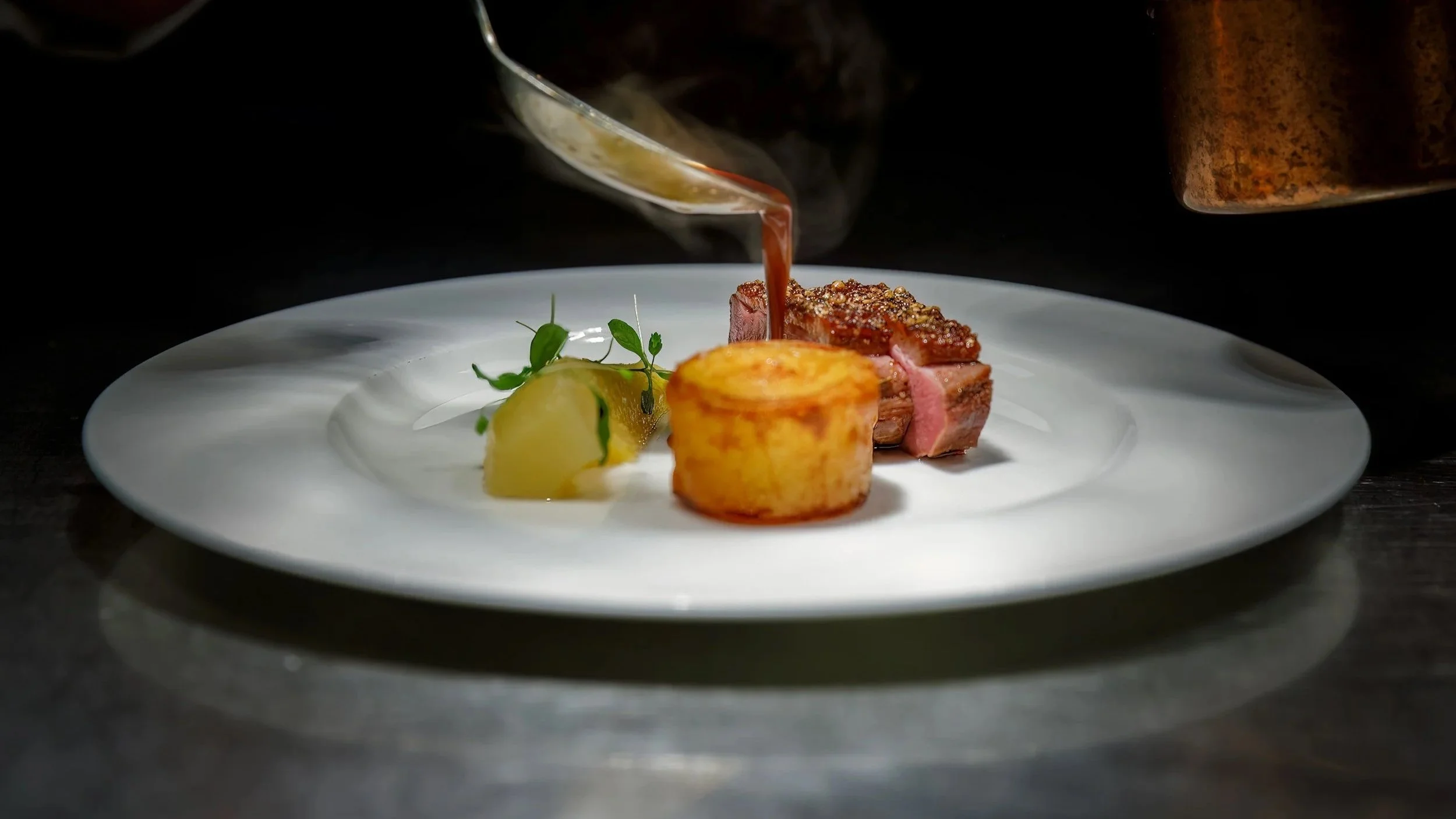 A fine dining plate with a piece of seared steak, a roasted potato, and a green garnish, with sauce being poured over the steak from a spoon.