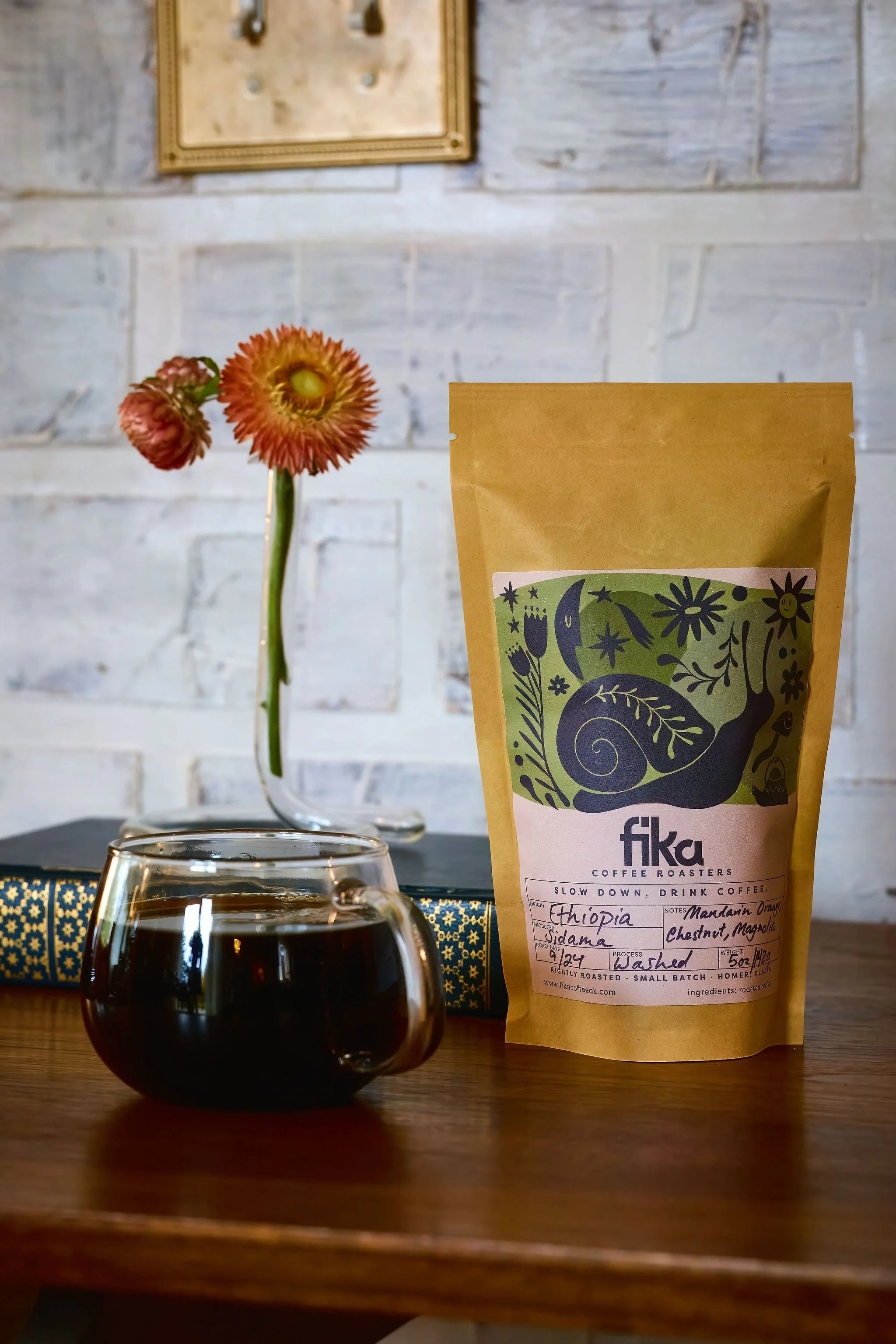 A bag of Fika Coffee Roasters Ethiopian Sidama coffee placed on a wooden table next to a glass of dark brewed coffee. In the background, a single pink and orange flower in a glass vase and a closed decorative book are visible against a white brick wall.