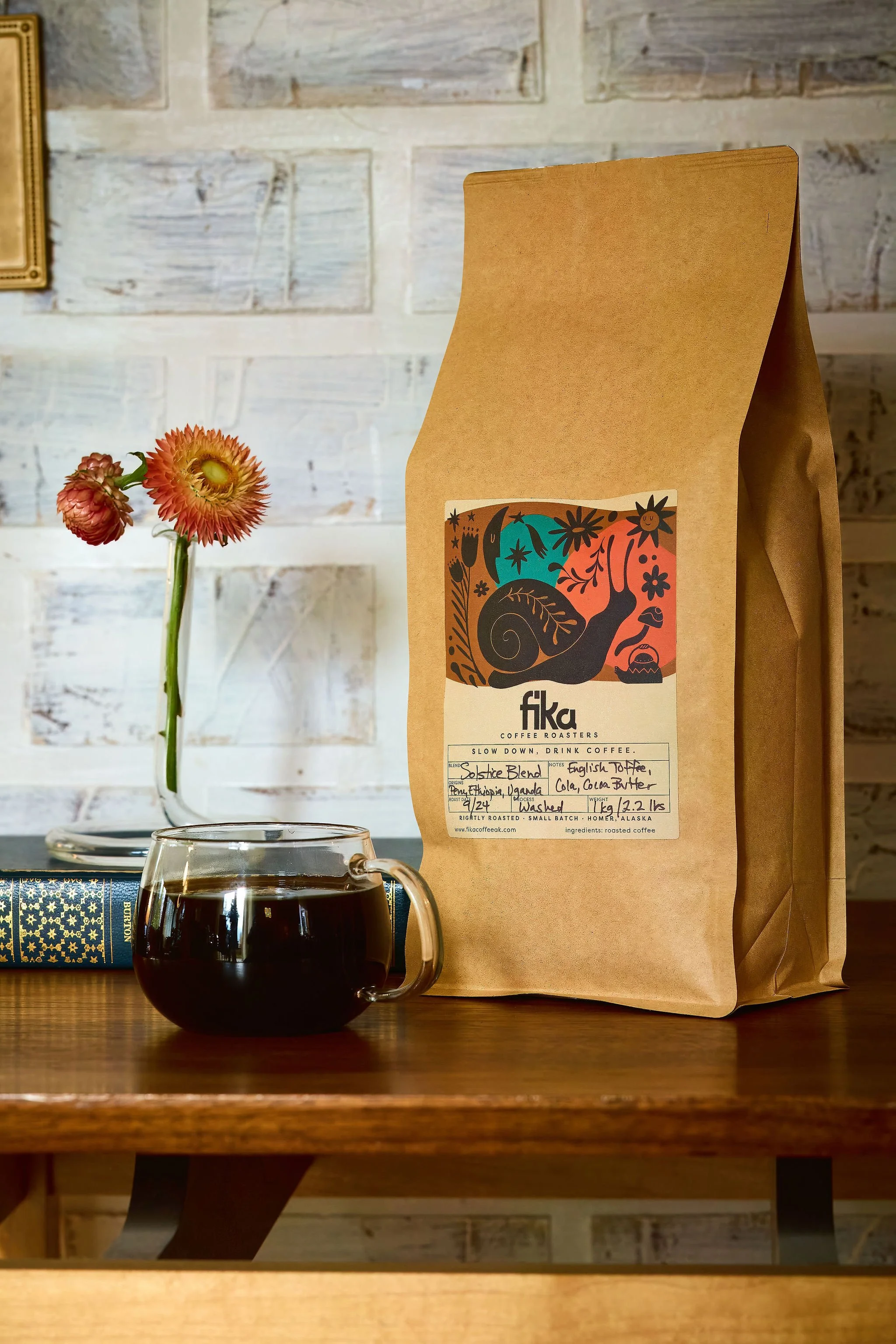 A brown paper bag of roasted coffee beans with a colorful label, a clear glass mug filled with black coffee, a small glass vase with pink and orange flowers, a closed hardcover book with a dark blue and gold cover, all on a wooden table against a white brick wall.