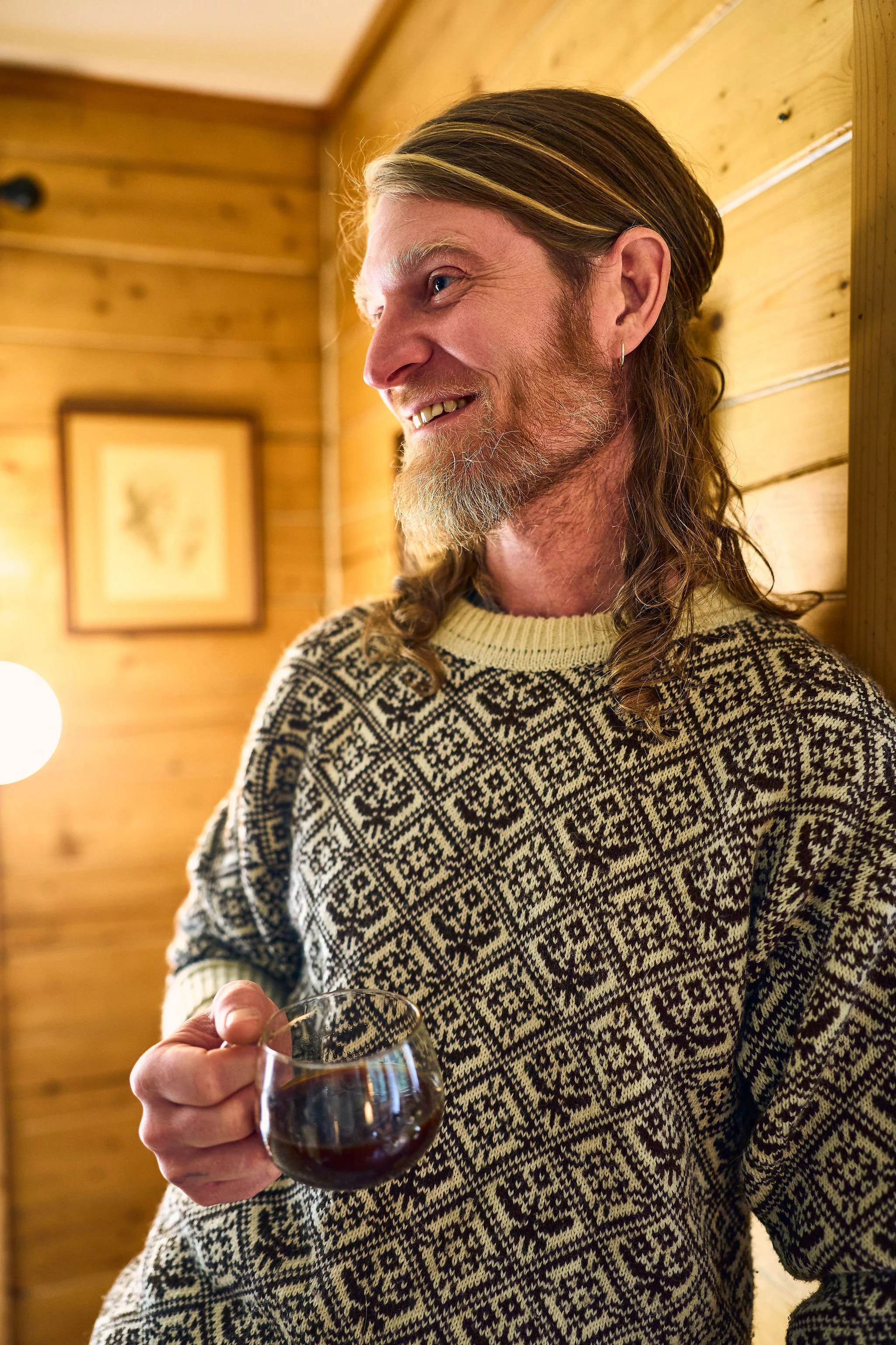 A man with long hair and a beard smiling while holding a glass of pour over coffee in a warmly lit wooden room.