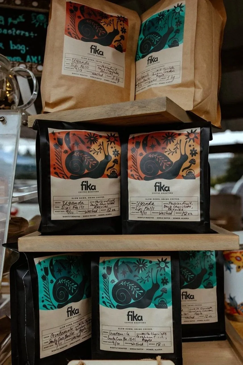 Multiple bags of Fika coffee with colorful, artistic packaging featuring a snail and floral designs, labeled with origin and roast details, displayed on wooden shelves.