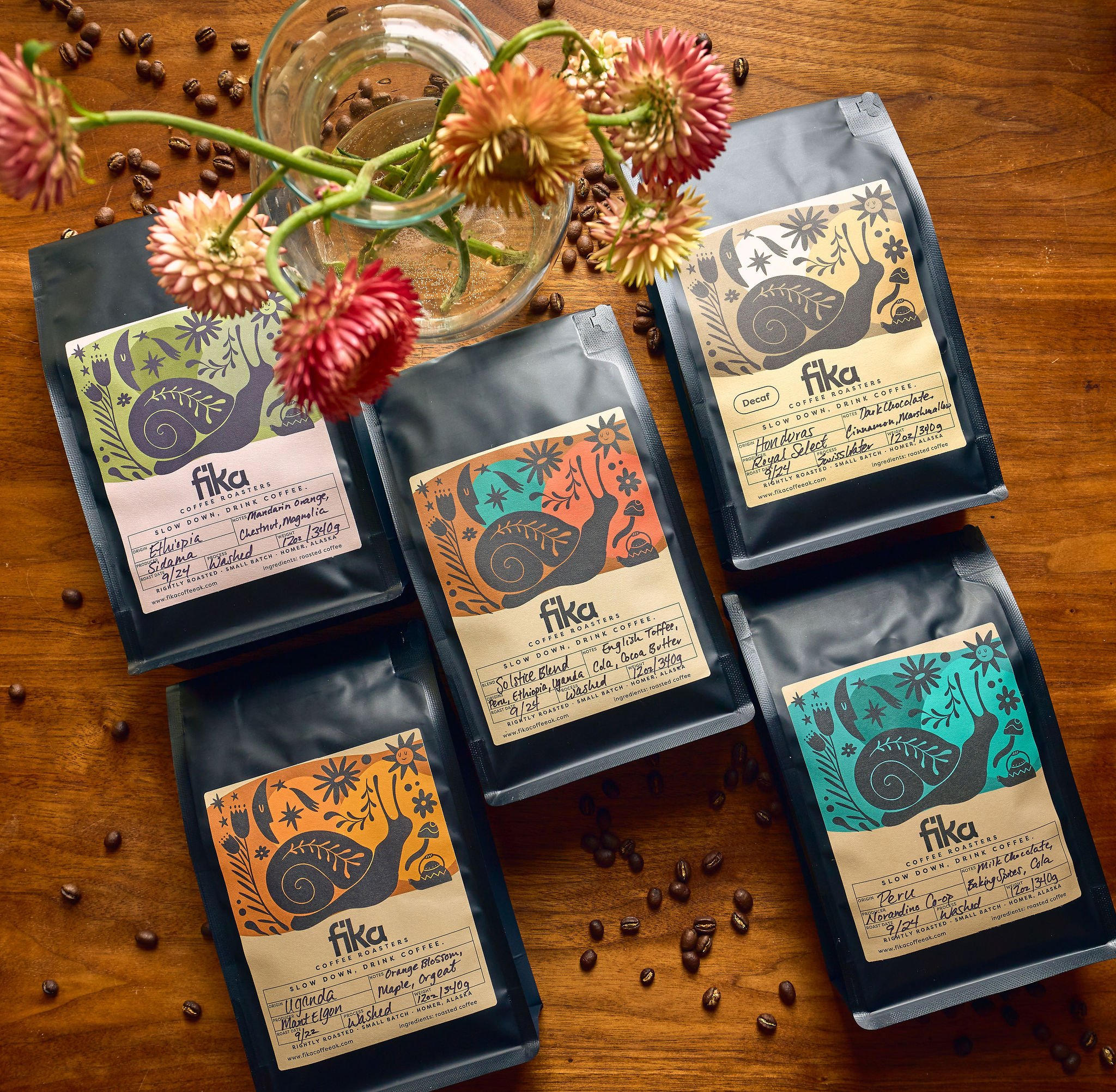 Five bags of Fika coffee with colorful labels, a glass vase with flowers, and scattered coffee beans on a wooden surface.