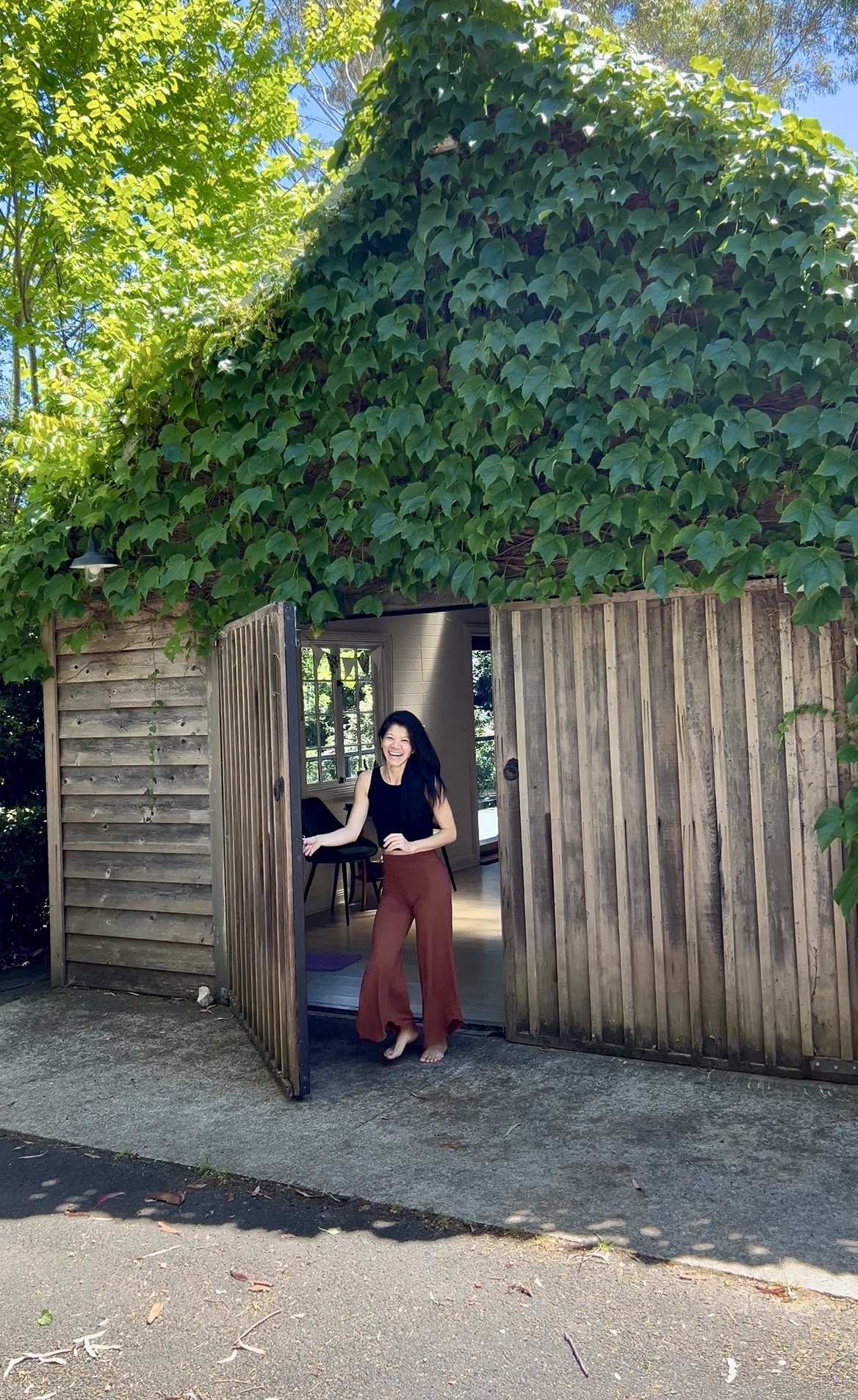 A woman standing in the doorway of a wooden shed, smiling. The shed is covered with large green ivy leaves and is open, revealing interior with a window and a chair. The surroundings include trees with green leaves and a paved area.
