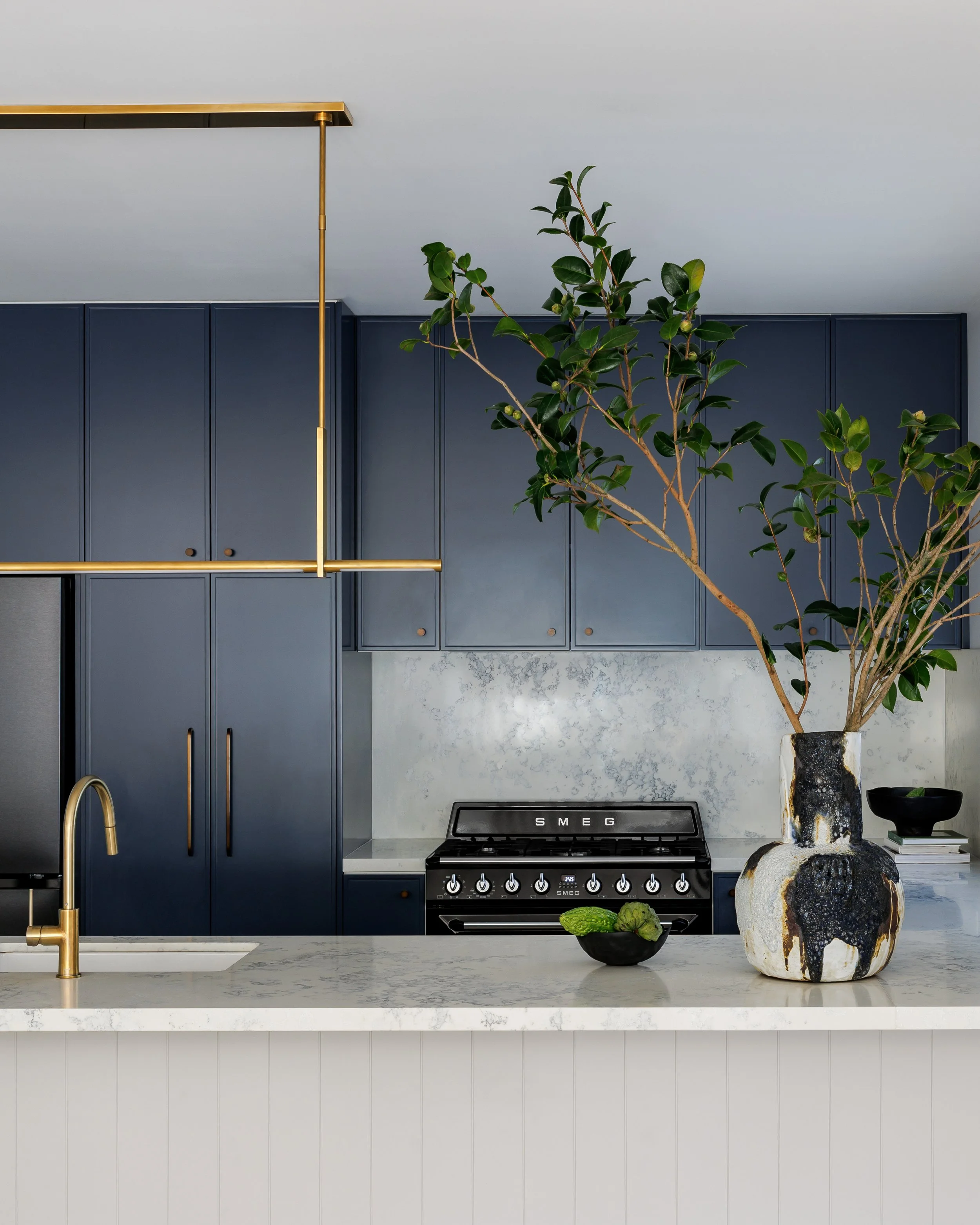 Modern kitchen with dark blue cabinets, white marble backsplash, marble countertop, gold faucet and fixtures, black stove, and decorative vases with greenery.