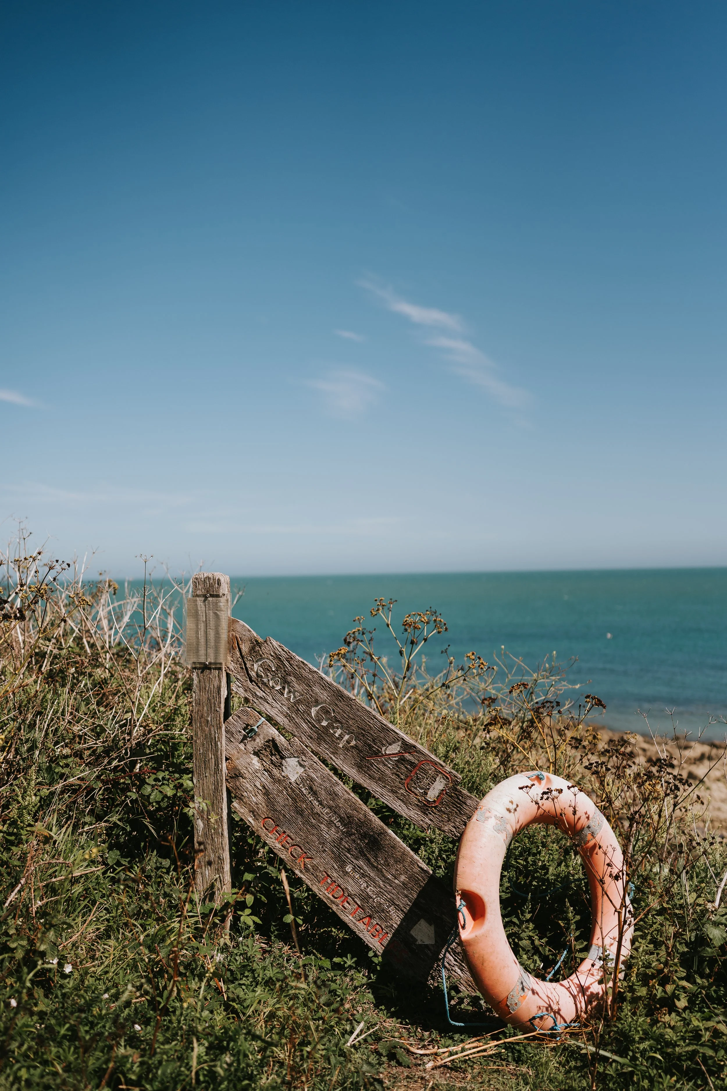 A faded wooden sign that says "Cow Gap" with a pink lifebuoy leaning on it. Blue skies and sea are visible in the background.