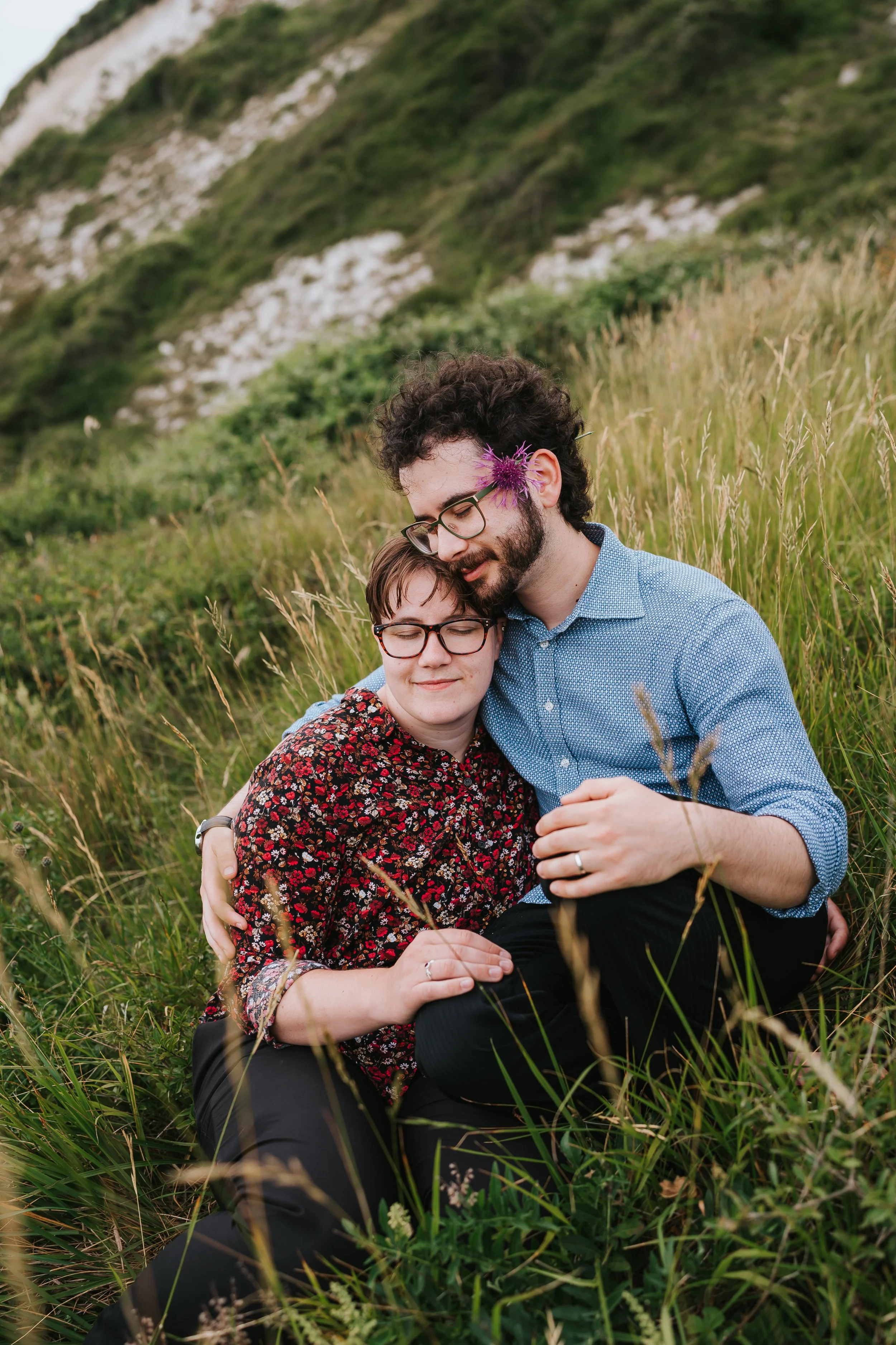 Brigid and Isaac holding each other close as they sit in the grass. Isaac has the purple flower over his ear.