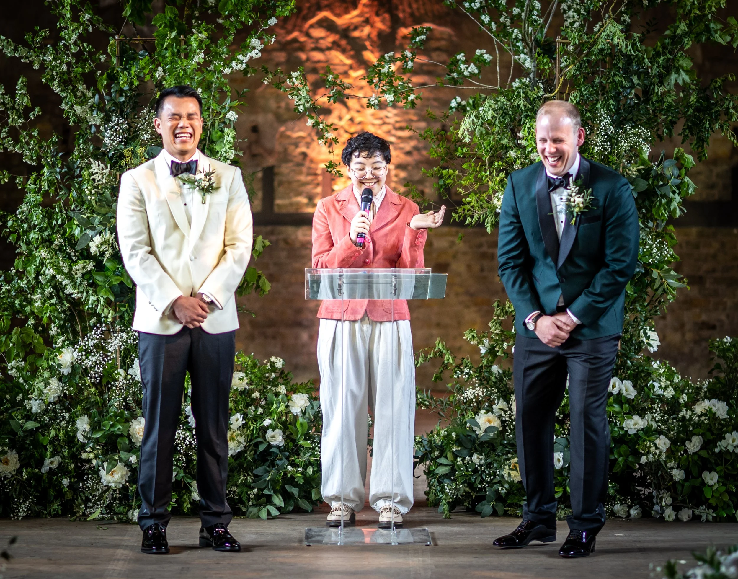 Two grooms, one is East Asian and in a white tux, one is white and in a dark green tux, laughing as Holden their celebrant, who is East Asian and wearing a pink jacket, stands between them and speaks.
