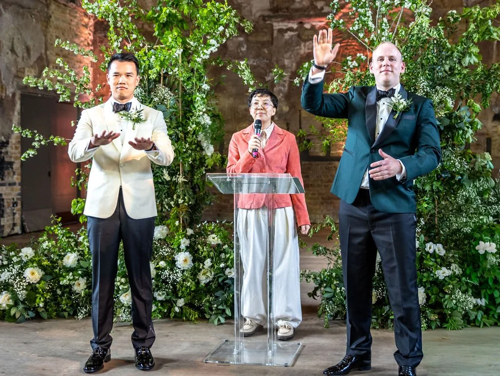 Kevin gesturing to tell guests to put their hands down while James is gesturing to tell guests to put their hands up for him. Holden their celebrant stands in between, looking at the results with amusement.