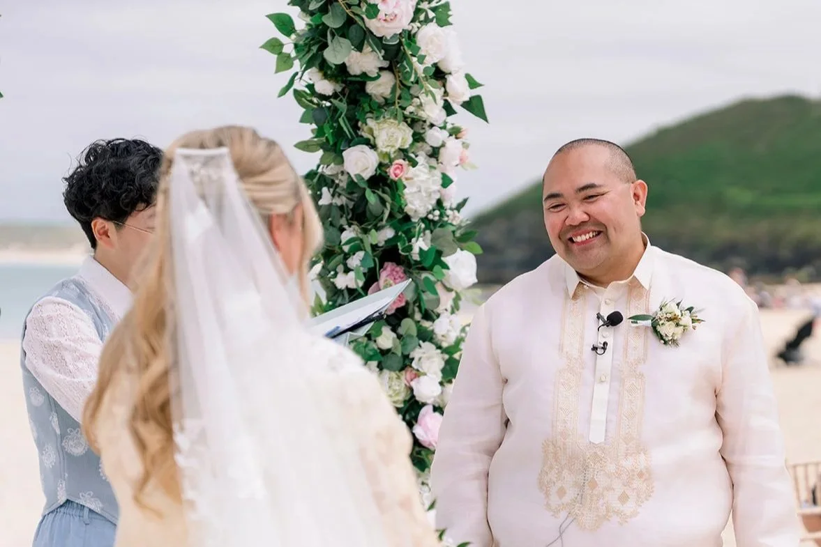 A beach wedding ceremony with a bride (her back is to the camera, she has long blonde hair and a white veil) and a groom who is Filipino. He is wearing a barong tagalog and smiling.