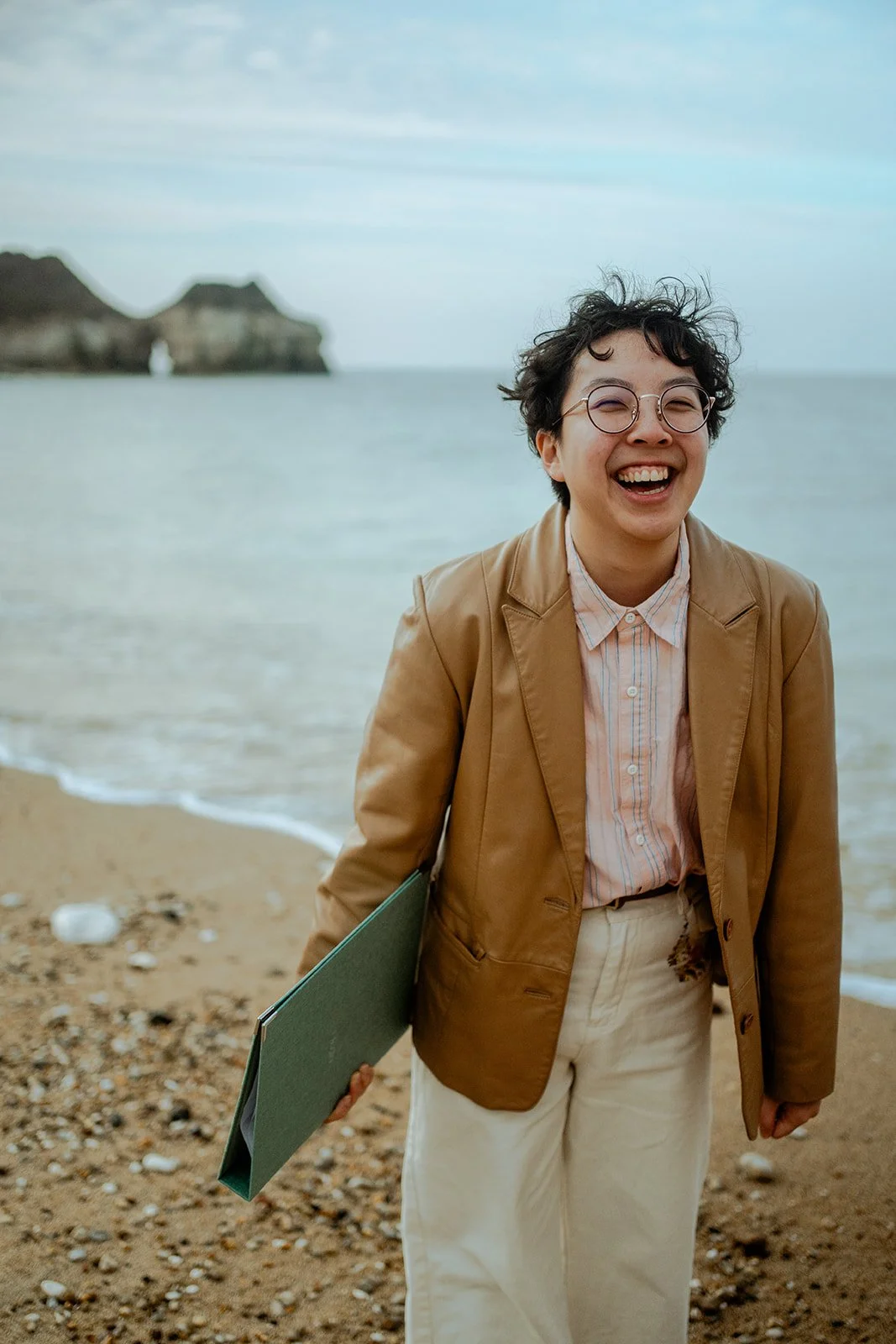Holden So, a wedding celebrant, laughing while standing on a beach. Holden is an East Asian nonbinary person with short black curly hair. They are wearing a beige leather blazer and holding a green ceremony folder.