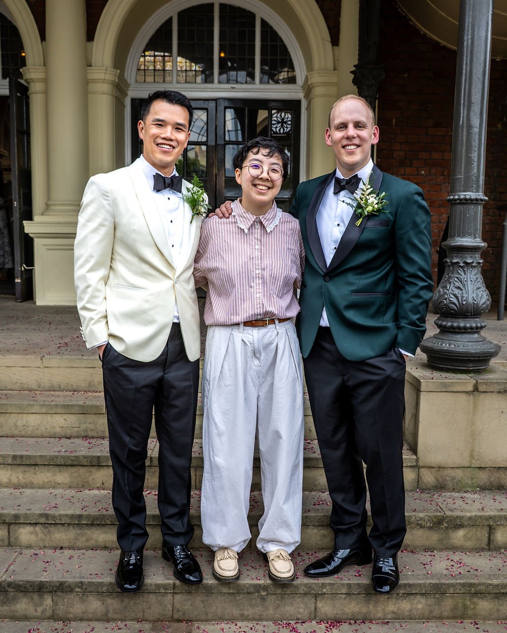 Kevin and James posing for a photo with Holden their celebrant, who has taken off the pink jacket to reval a striped pink shirt with a frilly collar.