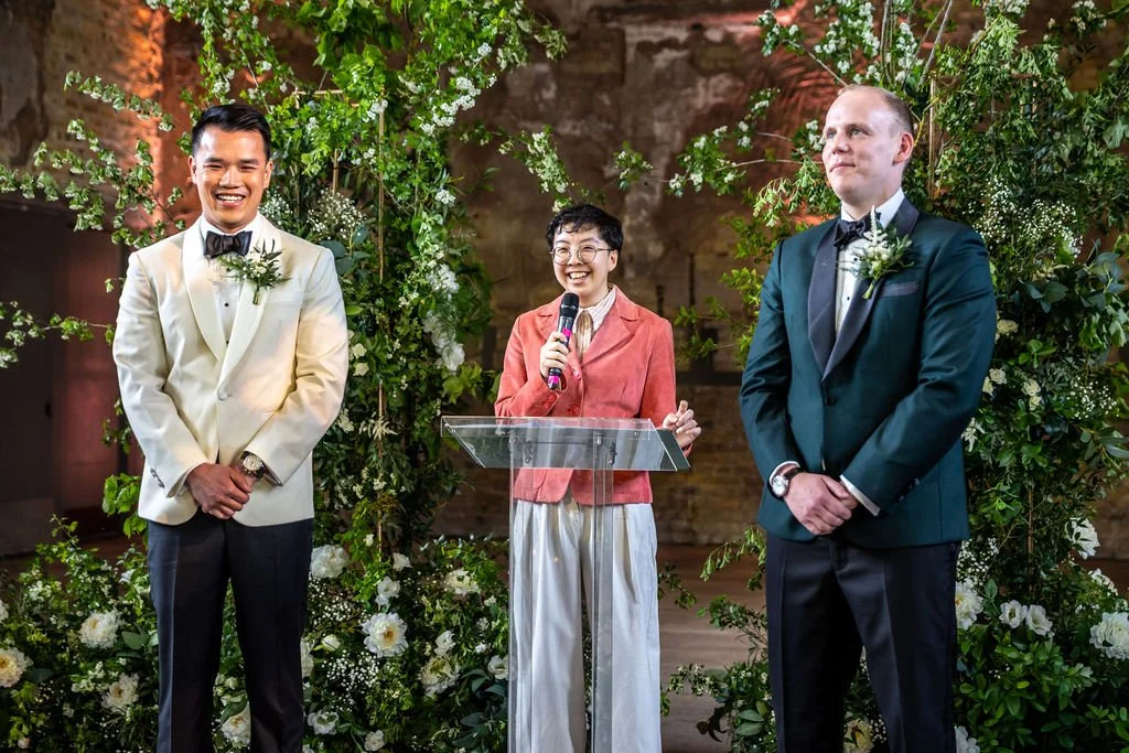 Kevin and James standing with their hands clasped while Holden their celebrant stands in between them and speaks. Holden is an East Asian nonbinary person and wearing a pink jacket.