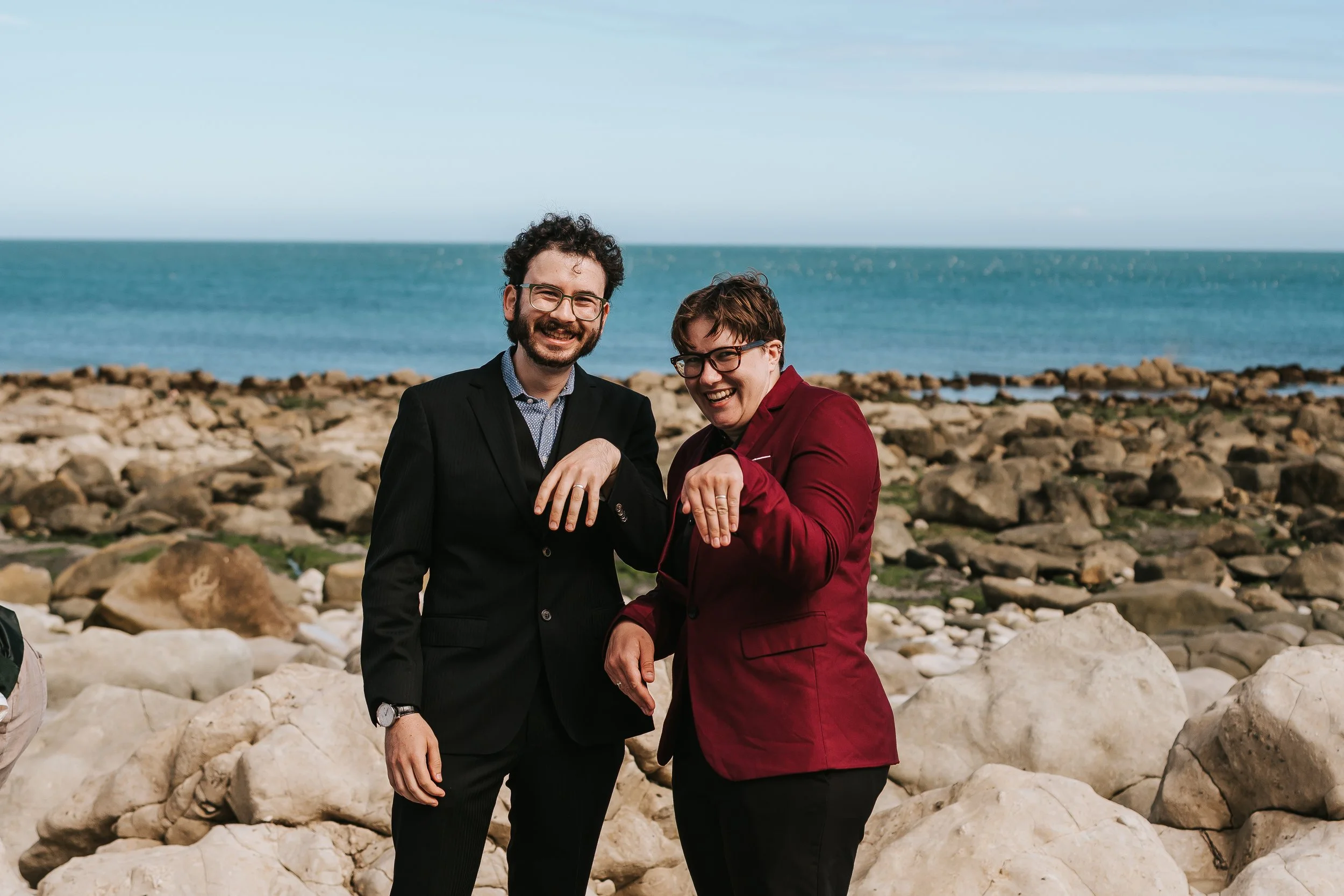 Brigid and Isaac showing off their wedding rings by holding up their hands. The sea and rocky beach visible in the background.