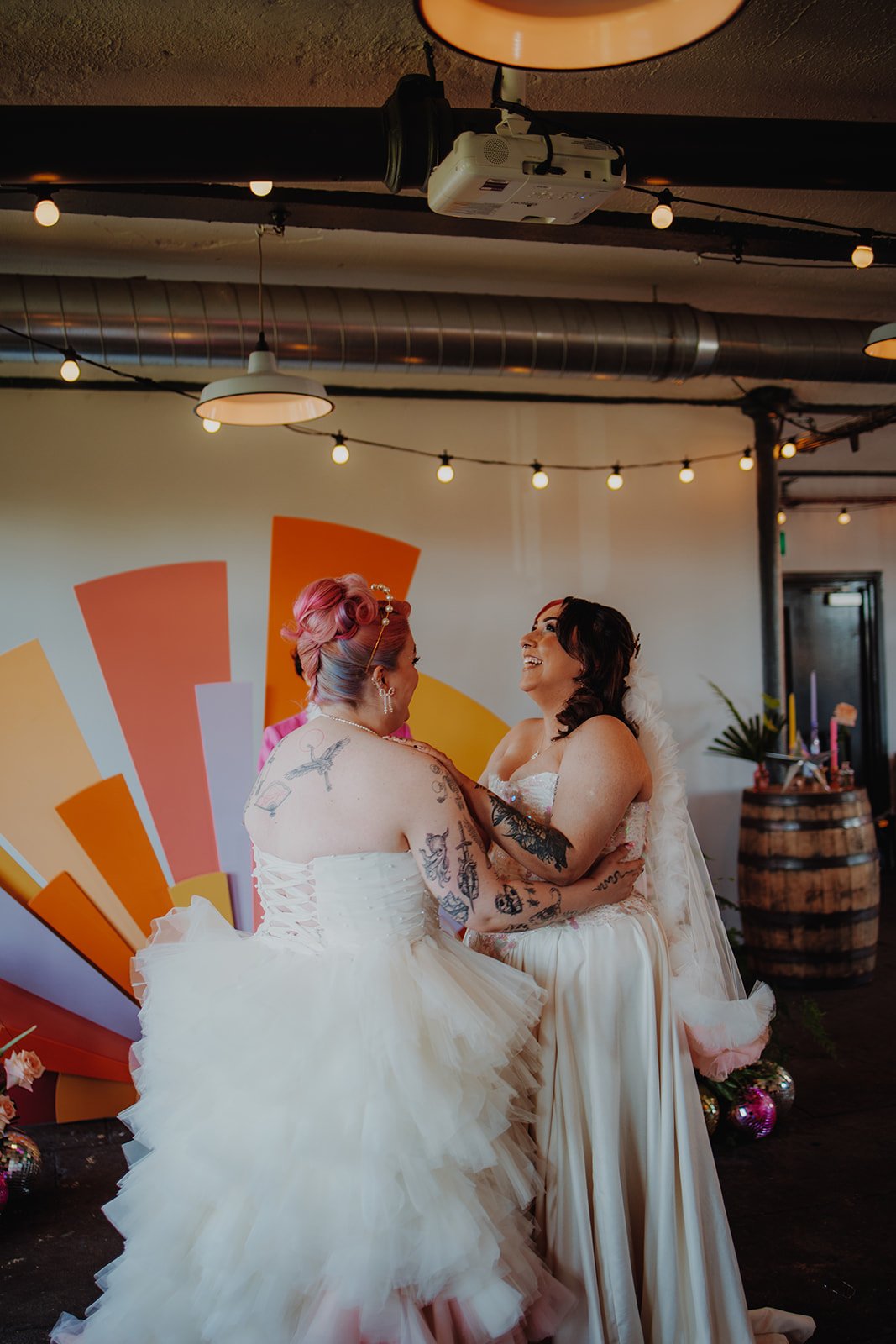 Two brides in white dresses - one is throwing her head back laughing. They both have visible tattoos.