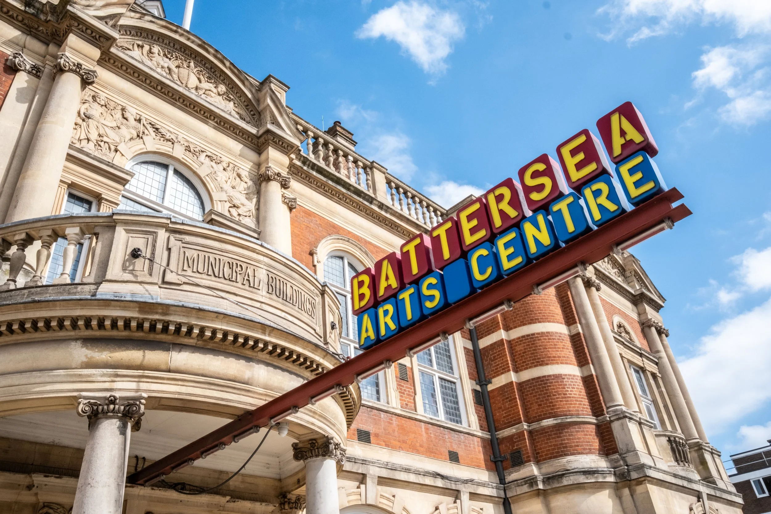 A red and blue sign that says "Battersea Arts Centre" with the letters in yellow, jutting outside a grand Victorian building.