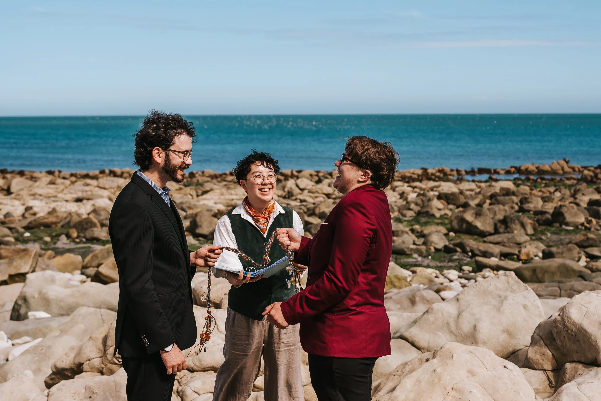 A beach elopement with a couple dressed in suits (one in black and one in burgundy) - both have short brown hair and wearing glasses. Their celebrant Holden is helping them perform a handfasting; all three of them have big smiles on their faces.