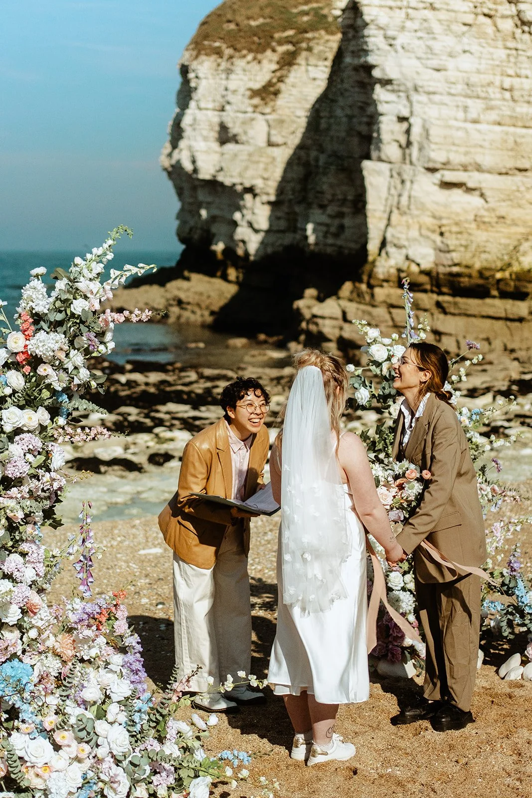 Holden So, an East Asian nonbinary celebrant, at a beach elopement with two brides, all laughing together