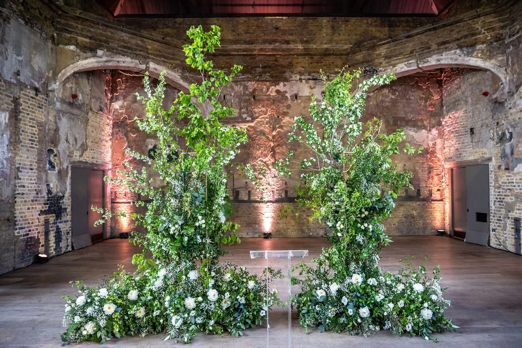 An broken floral arch that is mostly green with a profusion of white flowers at the base. Behind are brick walls with plasterwork that again looks damaged by fire.
