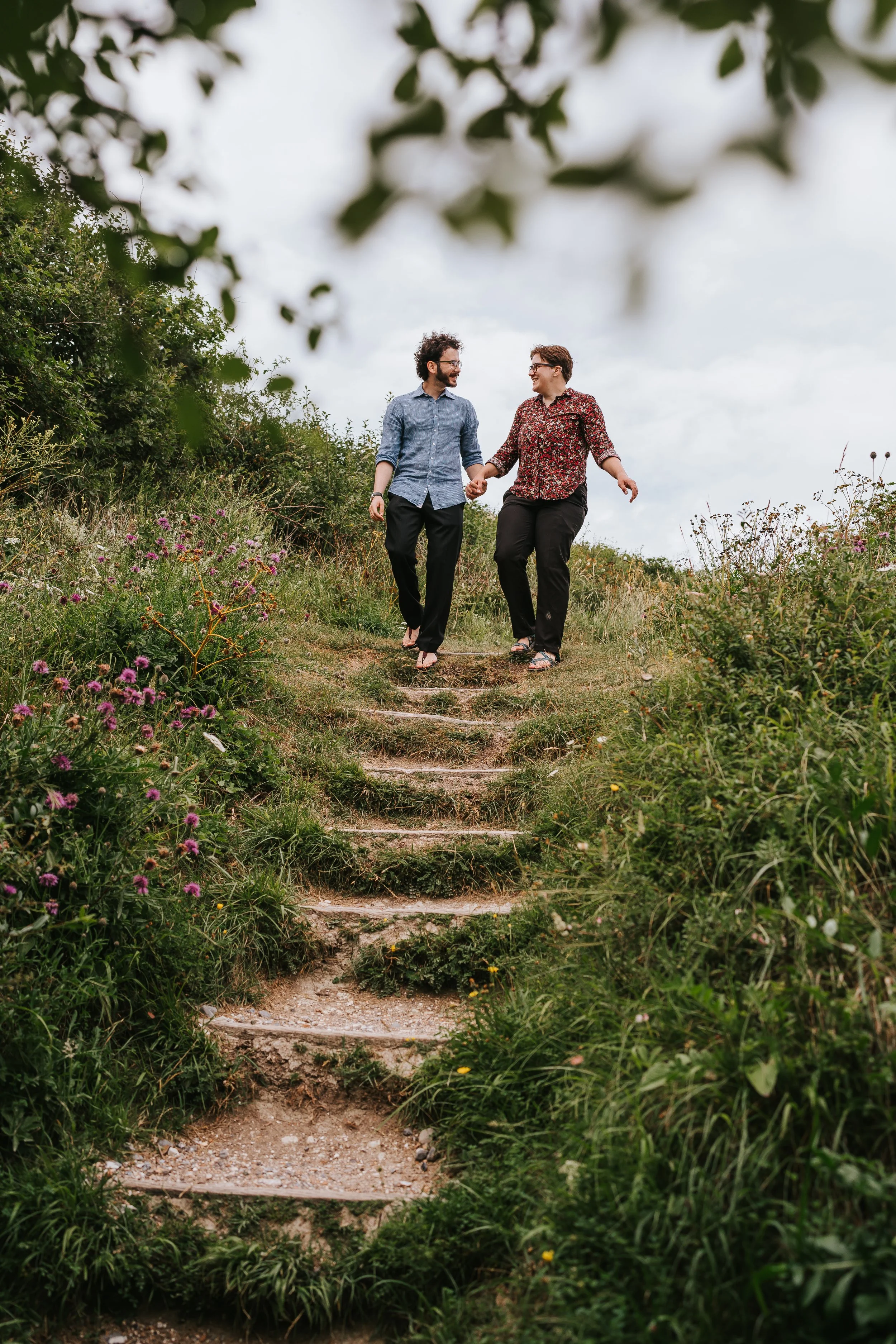 Brigid and Isaac walking down some steps with greenery all around them, and pink wildflowers.