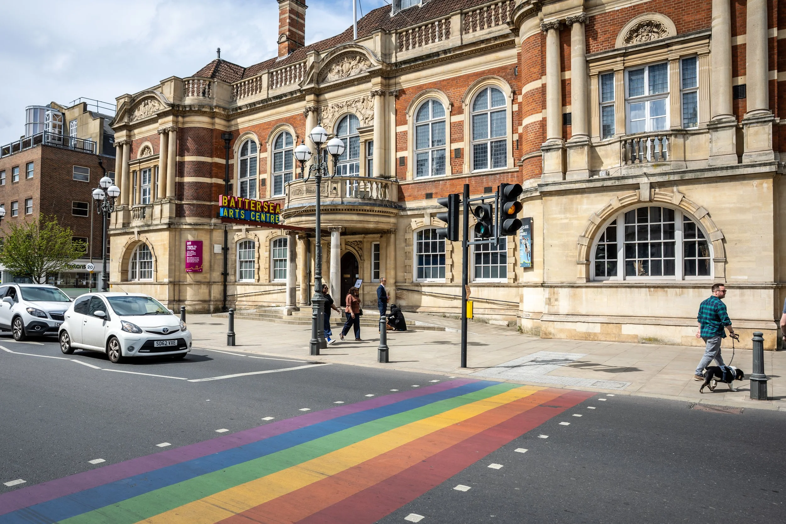 A traffic crossing outside the Battersea Arts Centre, a grand-looking Victorian building. The ground of the traffic crossing has been painted in the colours of the rainbow flag.