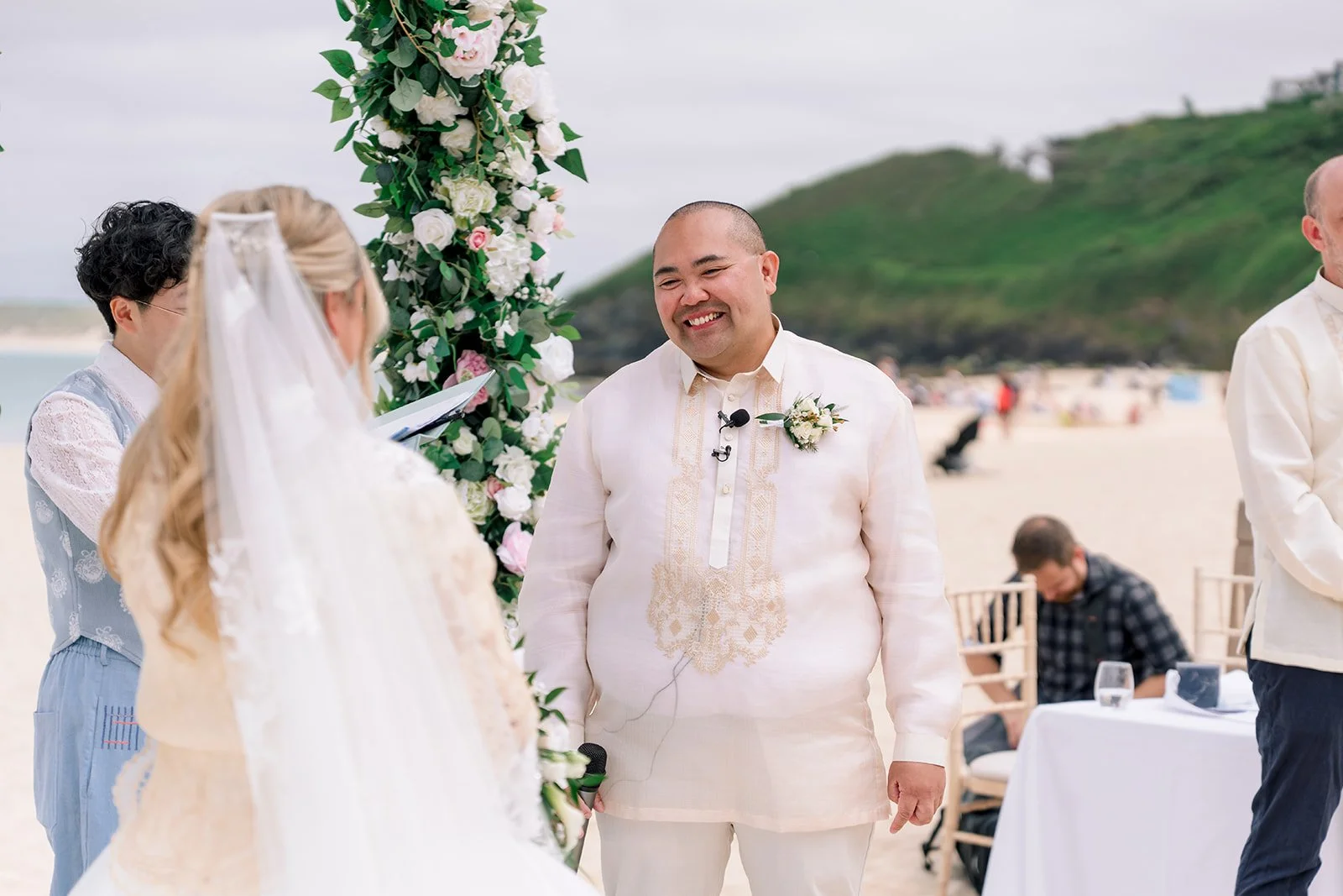 A wedding ceremony on a beach. The bride's back is to the camera, her long blonde hair and white veil are visible. The groom is facing the camera and smiling; he is Filipino and wearing a barong tagalog.