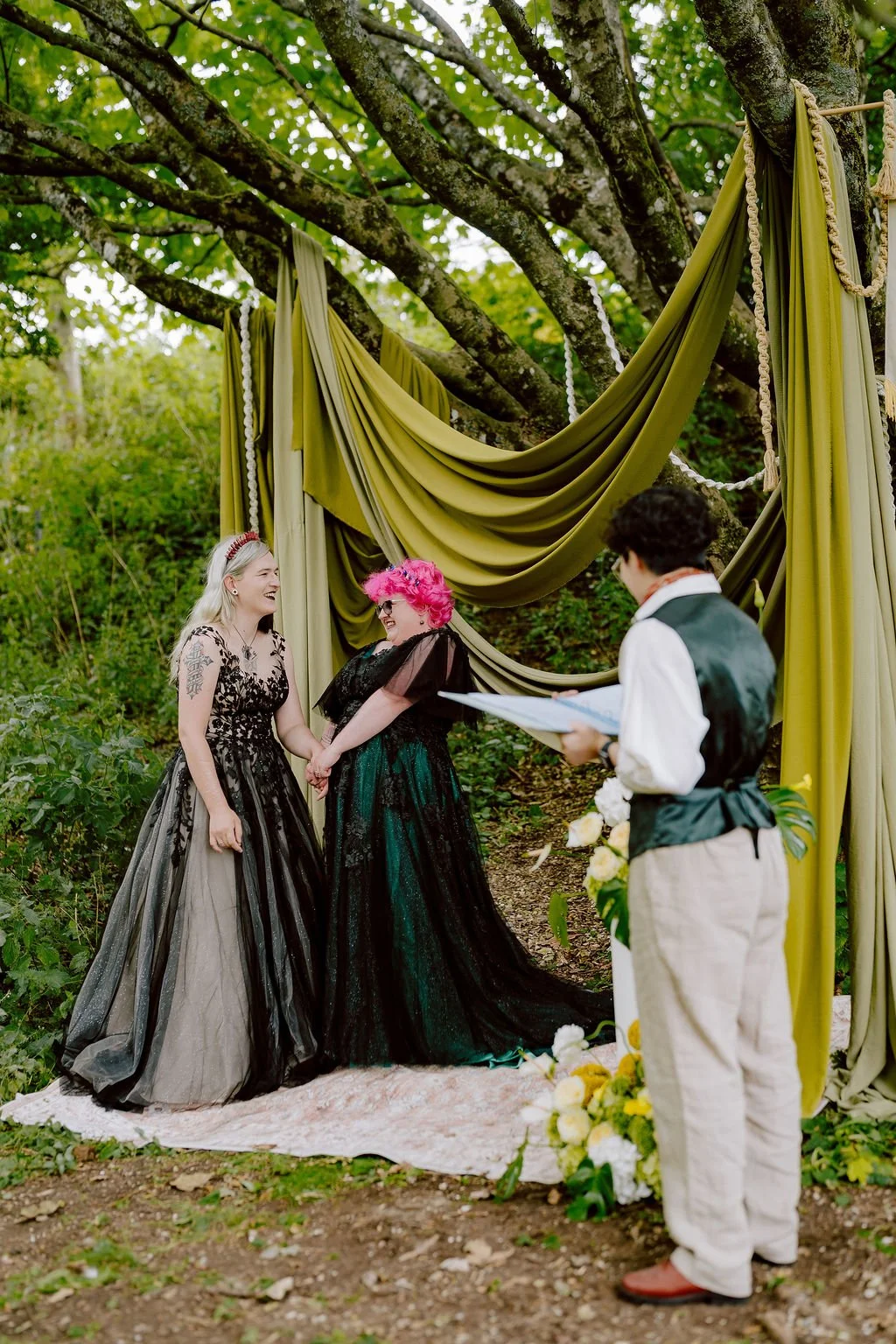 Alexis and Zoe with their celebrant Holden So. Alexis and Zoe are both white women; Alexis has long blonde hair and Zoe has short pink hair and they are both wearing dark dresses.