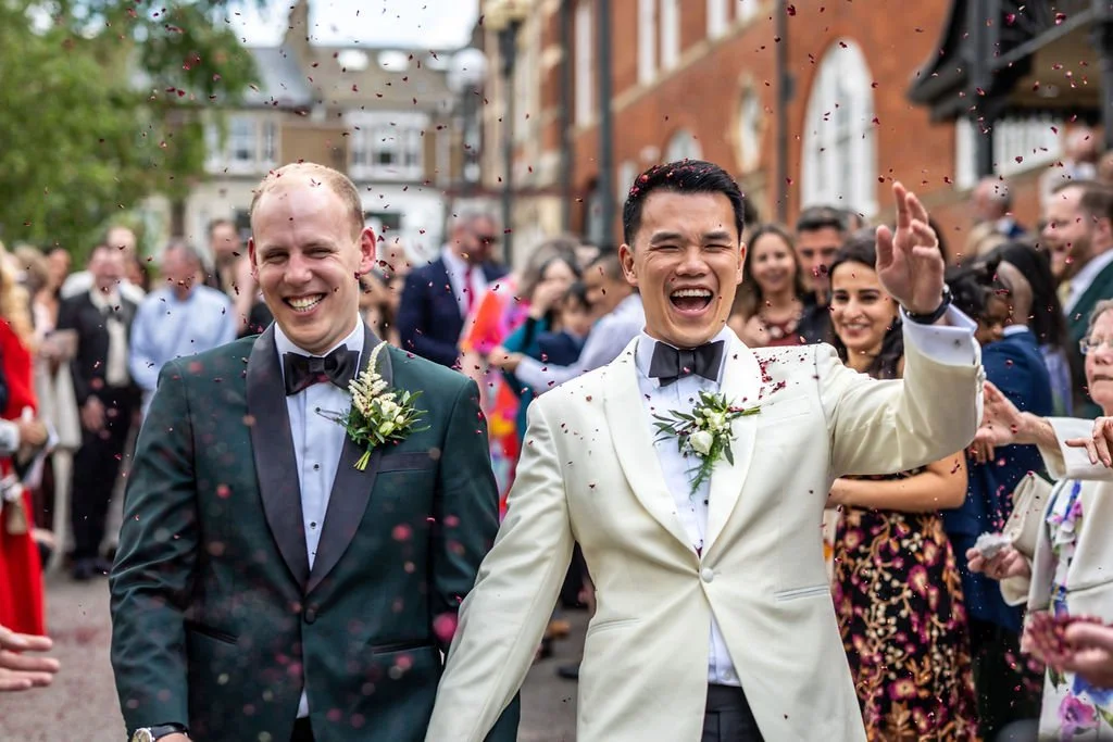 Kevin and James laughing while confetti in the form of dark pink petals rain down on them. Their guests stand around them.