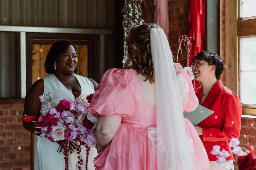A bride in white smiling at another bride in pink, while Holden leads their wedding ceremony