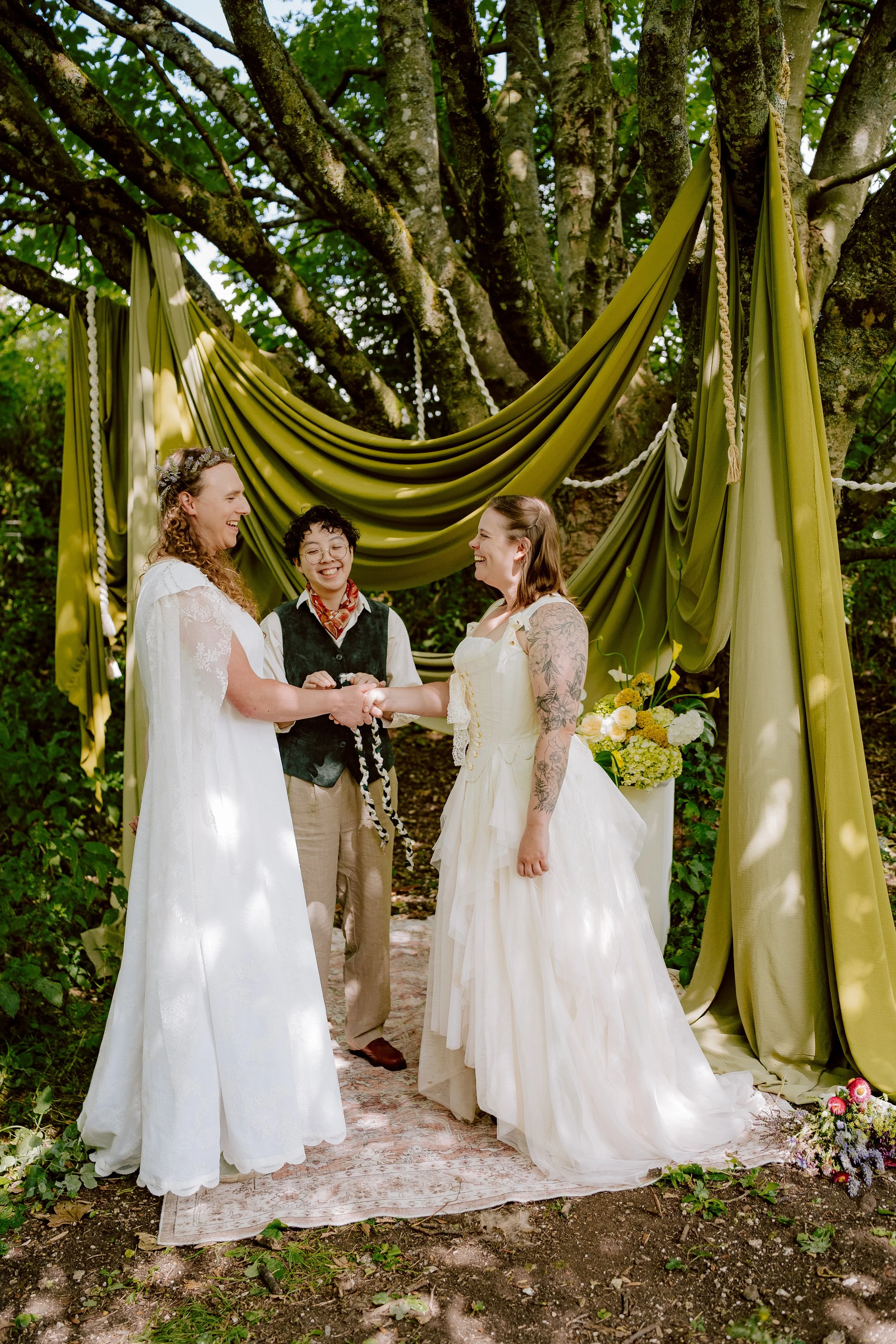 Two women in white dresses smiling at each other as Holden their celebrant is performing a handfasting for them with a braided cord.