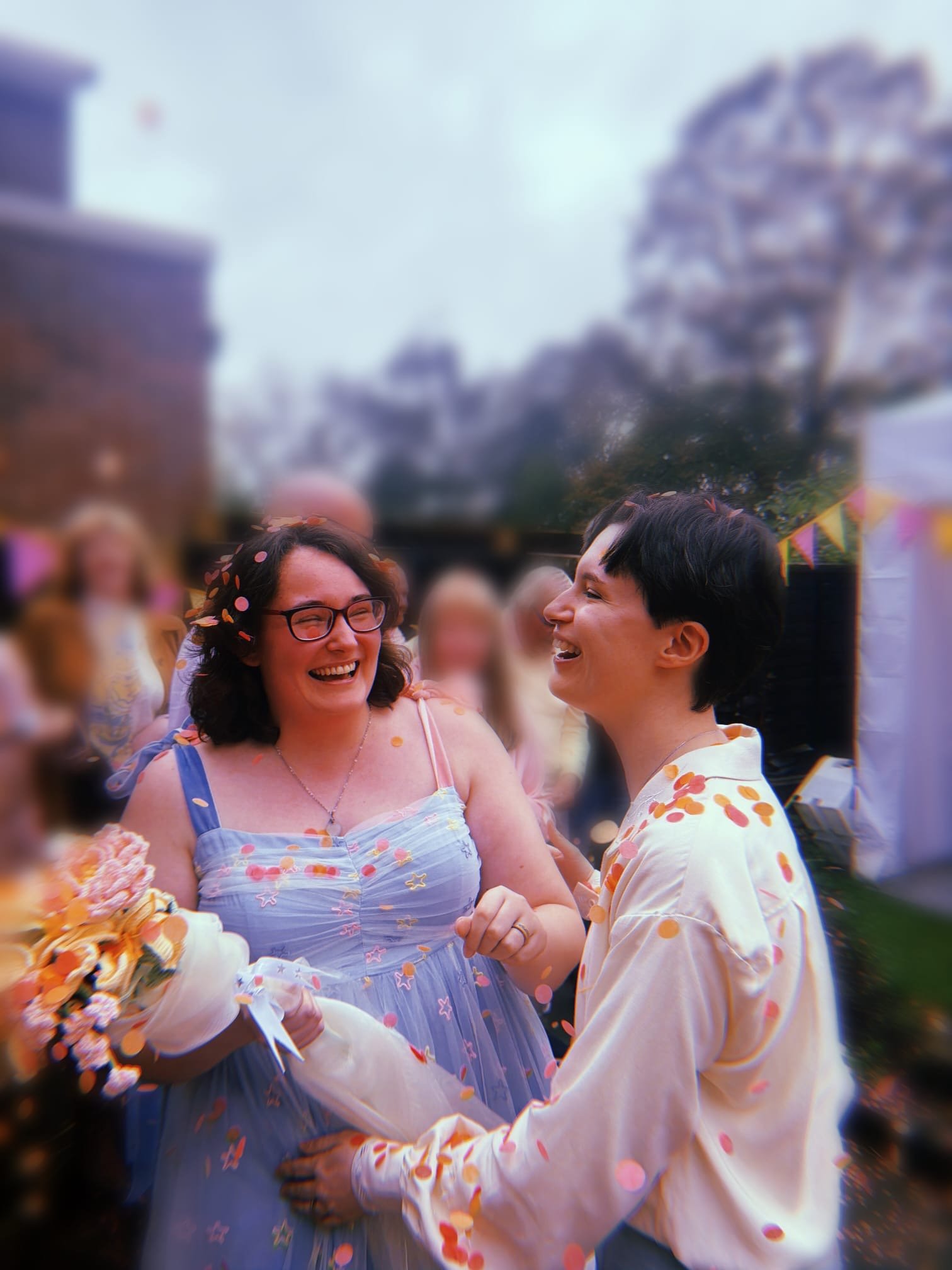 Adele and Tabby laughing and covered in pink and orange confetti. Adele has shoulder-length brown hair and is wearing a blue dress with one blue strap and one pink strap. Tabby has short brown hair and is wearing a pale coloured shirt.