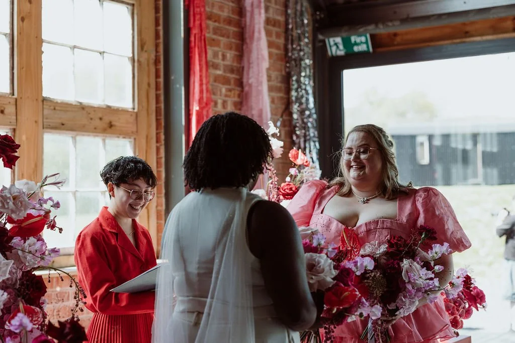 Yolly and Nikki standing with their celebrant Holden So. Yolly is a white woman in a pink dress and Nikki, who has her back to the viewer, is a Black woman in a white jumpsuit.