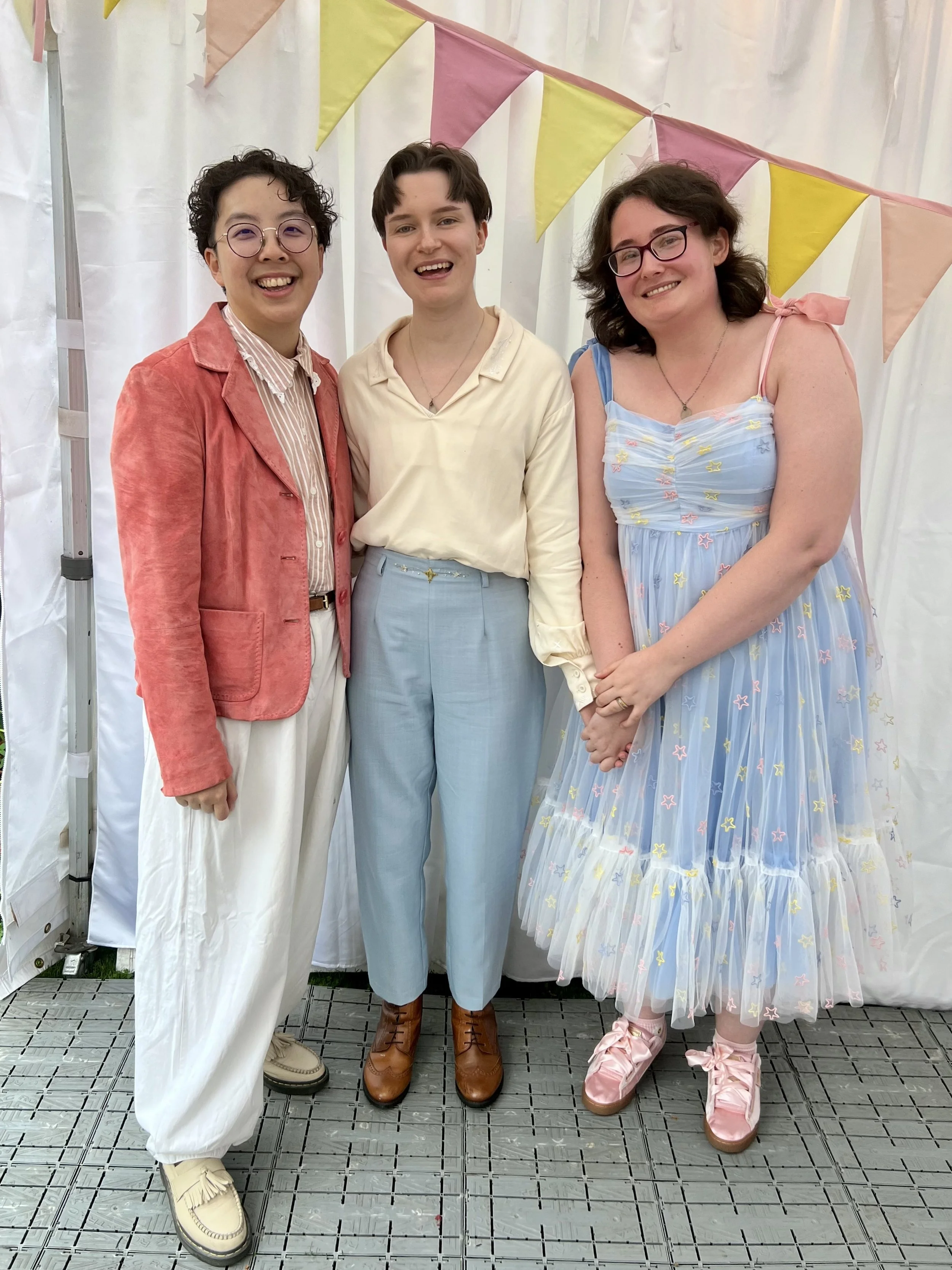 Tabby and Adele posing for a photo with their wedding celebrant, Holden. Holden is an East Asian person with short black hair, wearing a pink suede jacket and cream trousers.