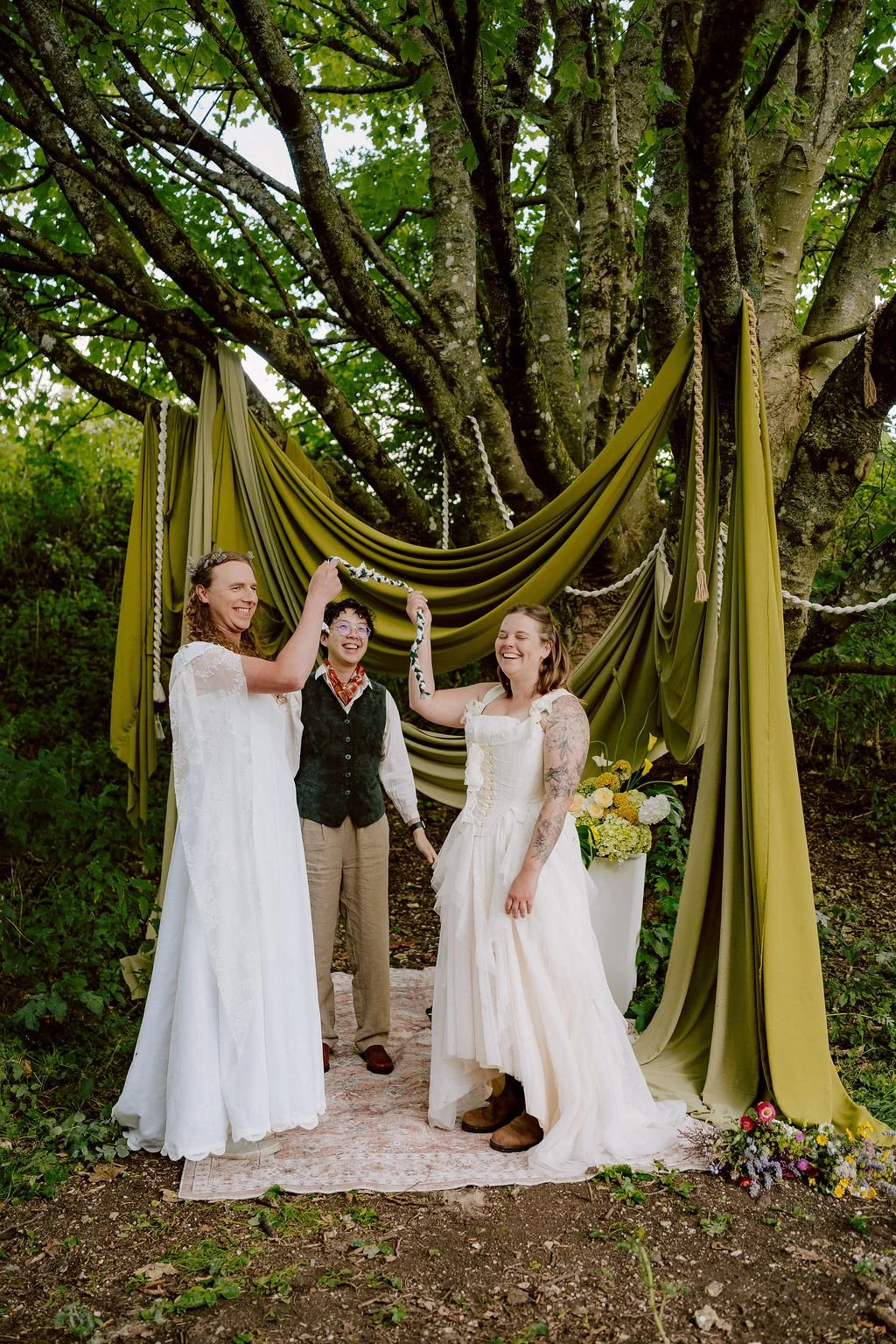 Elle and Myra with their celebrant Holden So. Elle and Myra are both white women wearing white dresses. They are holding up their handfasting cord and smiling.