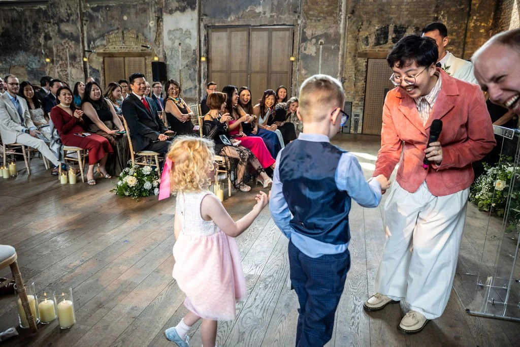 The backs of two children, one a girl in a pink dress and one a boy in a blue suit, as they hand over the rings to Holden the celebrant. Guests are all looking delighted by this adorable sight.