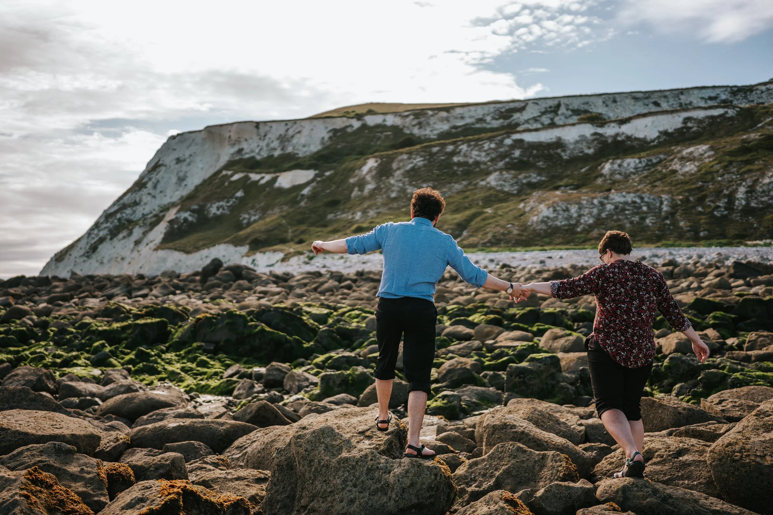 Brigid and Isaac with their back to the camera as they walk over big rocks while holding hands. Isaac is in a blue shirt and Brigid in a burgundy floral shirt. There are white cliffs in the distance.