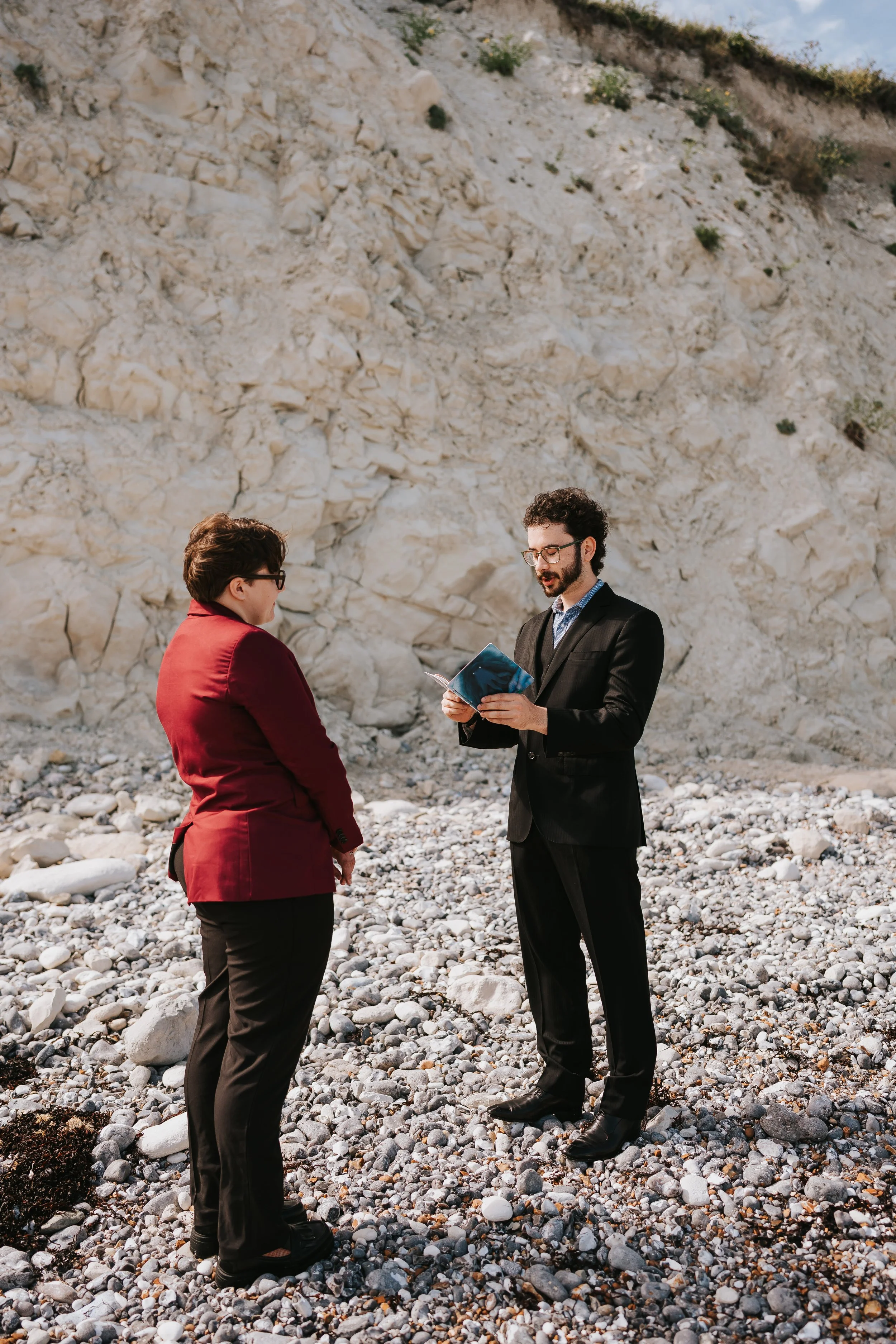 Isaac reading from a blue notebook while Brigid listens, with the white cliffs behind them.