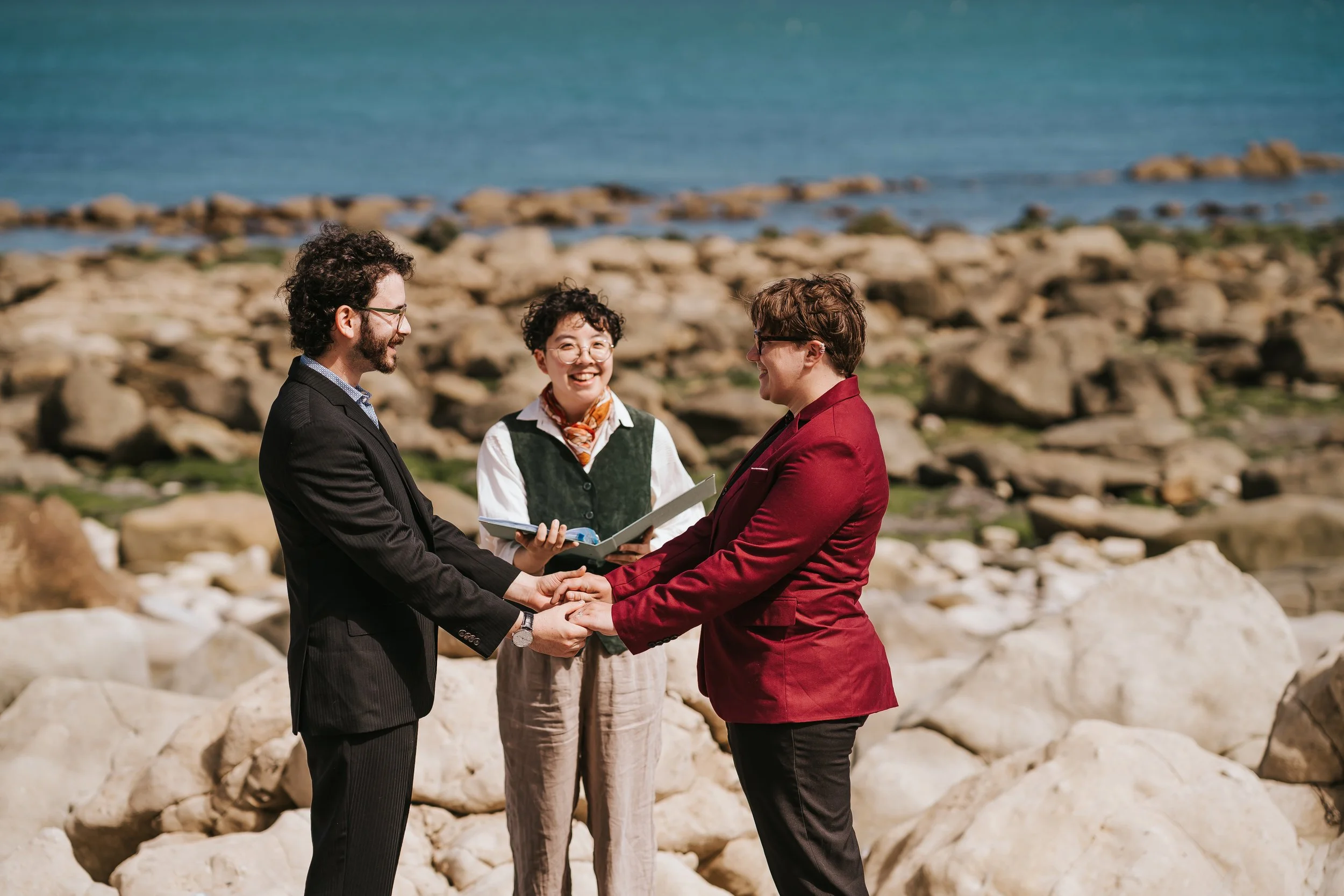 A closer shot of Brigid and Isaac during their beach elopement with Holden their celebrant standing between them. They are all smiling.