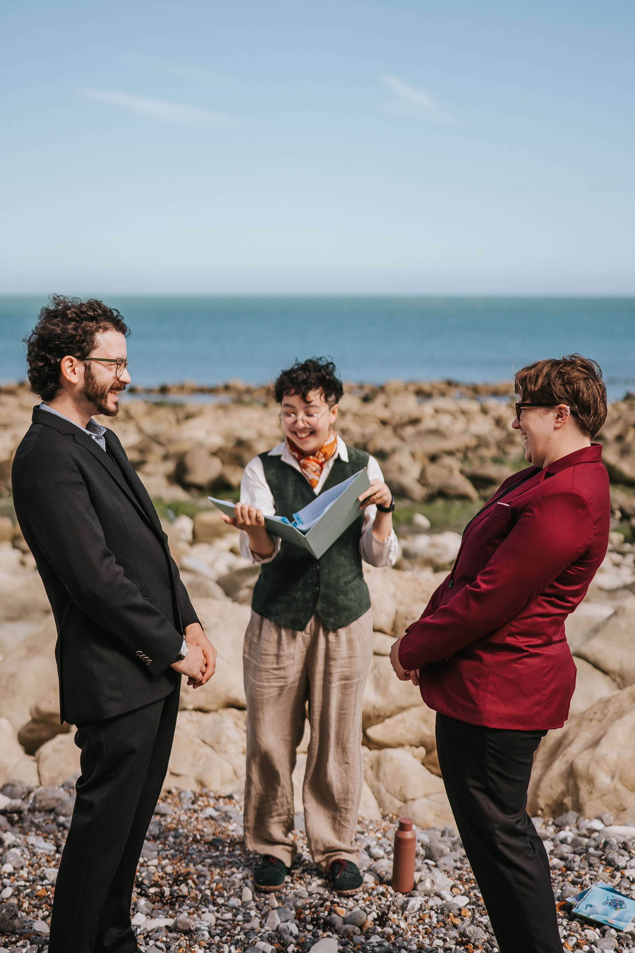 A beach elopement featuring a couple dressed in suits (one in black, one in burgundy). They are laughing with the sea in the background and Holden as their celebrant between them, dressed in a dark green waistcoat and beige trousers.