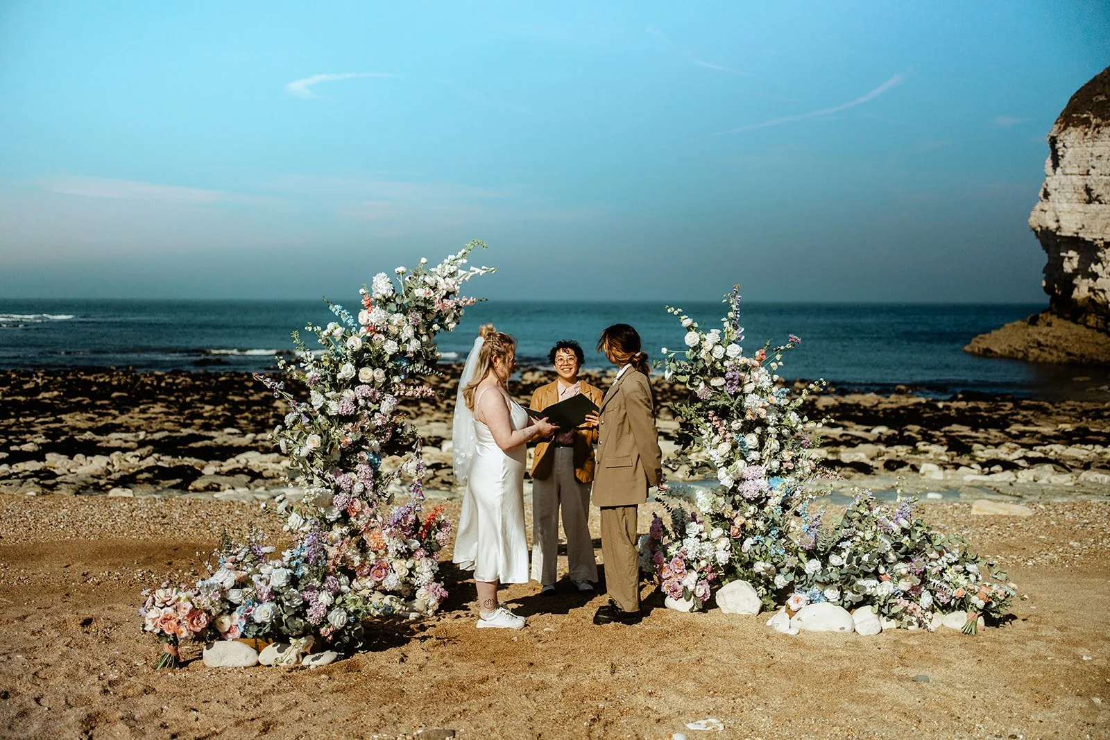Two brides, one in a white dress and the other in a khaki suit, standing on a beach in between a broken floral arch, with Holden So their celebrant between them.