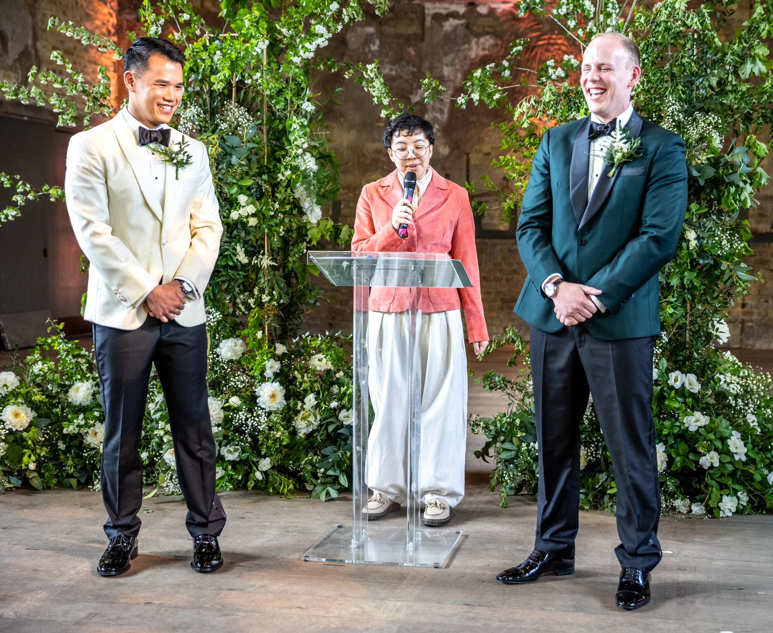 Kevin, an East Asian man in a white tux, and James, a white man in a pine green tux, laughing while Holden their celebrant is speaking. Holden is in a bubblegum pink suede jacket and white trousers.