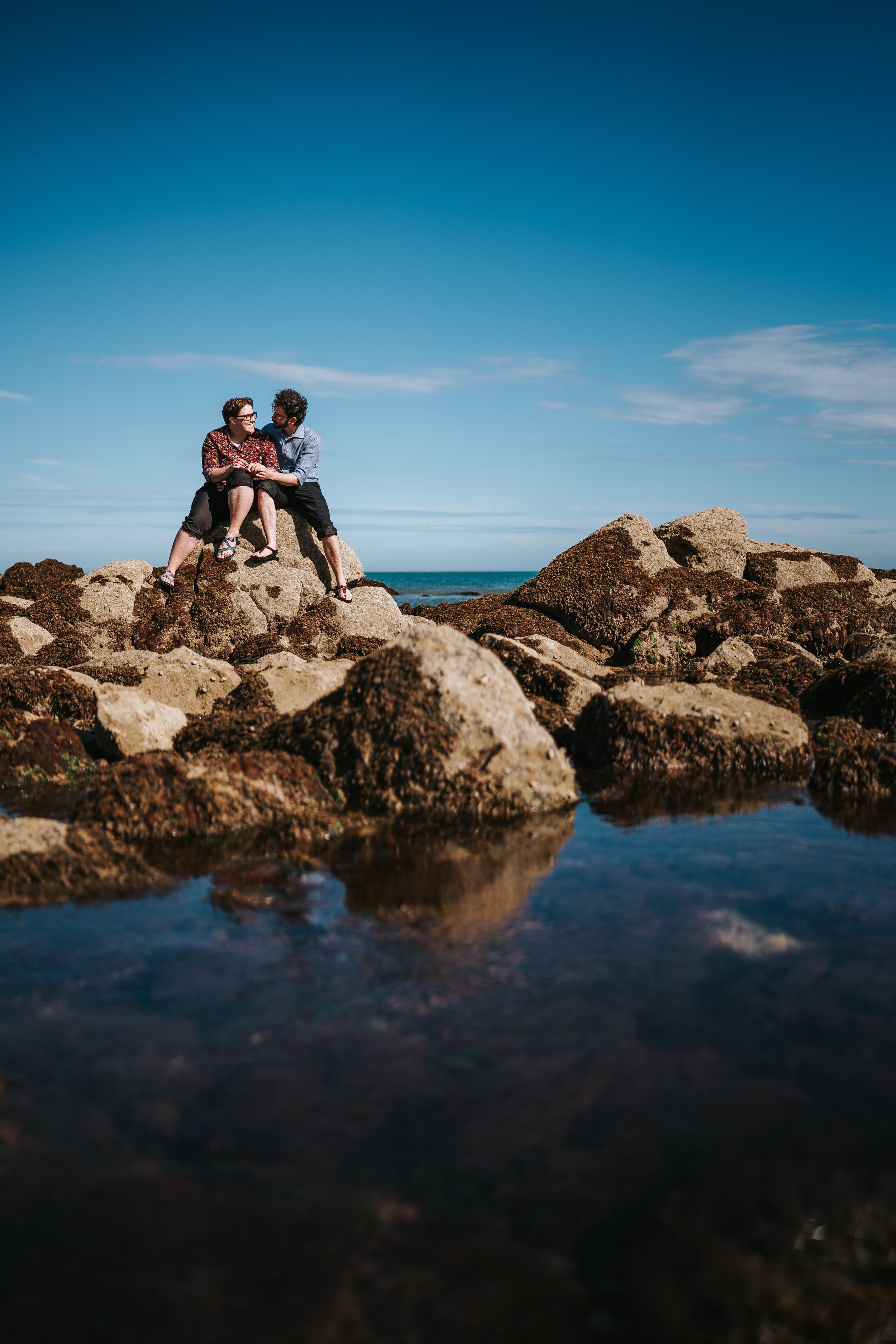 Brigid and Isaac sitting on a big rock, water below them and blue skies above.