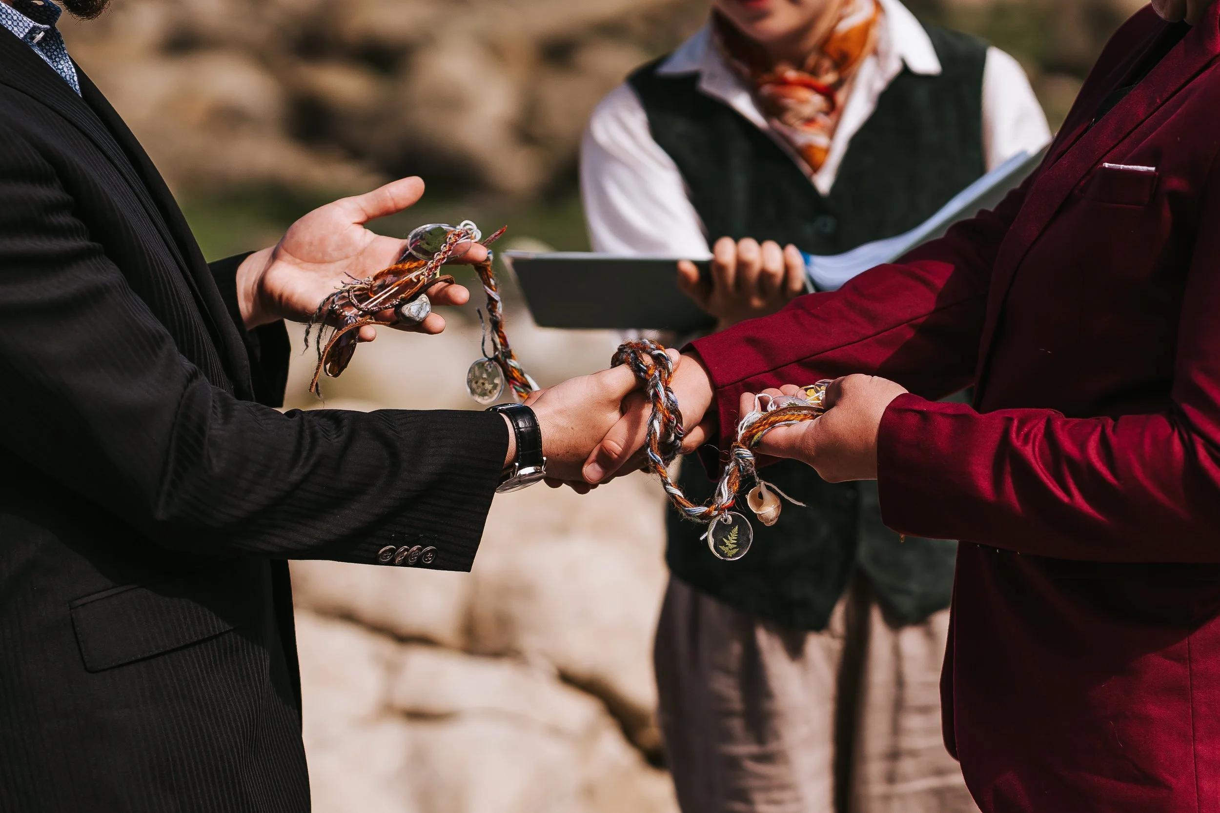 A close-up of Isaac and Brigid's hands as they perform the handfasting ritual. The handfasting cord is made of colourful strands of yarn and has shells and flower beads attached to it.