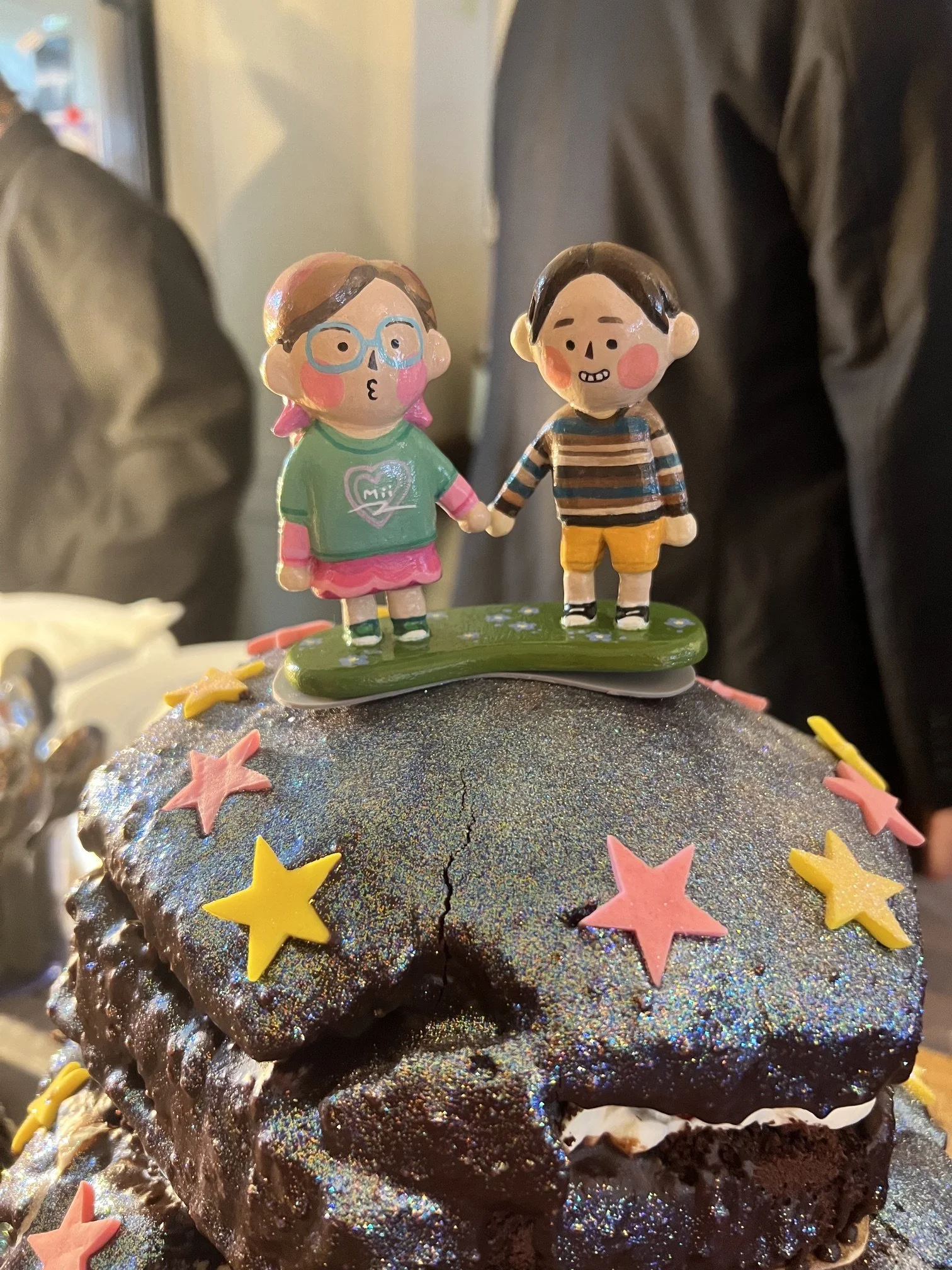 A colourful clay cake topper on top of a sparkly chocolate cake that has pink and yellow stars on it. The cake topper features Adele and Tabby holding hands and blushing.