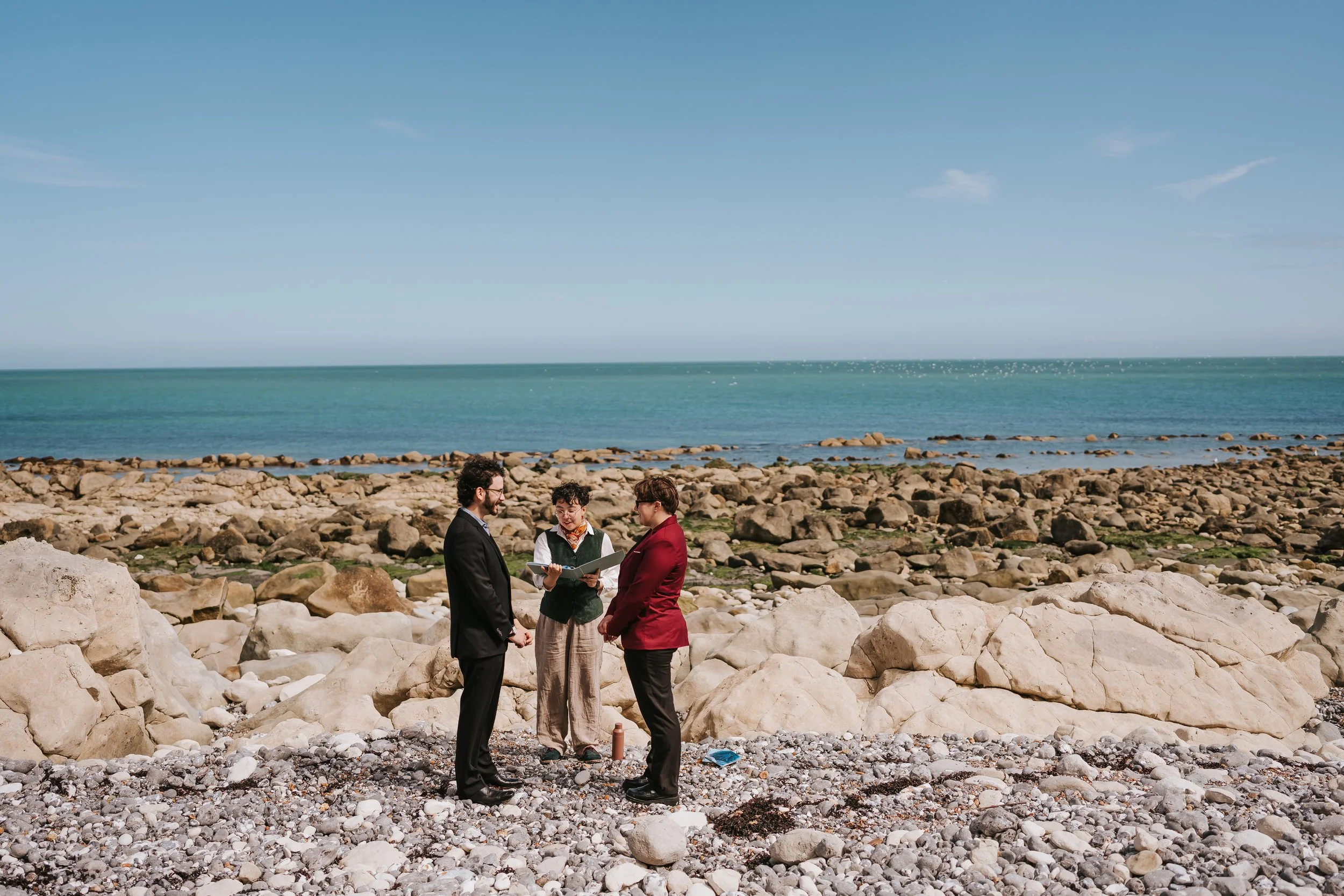 Brigid and Isaac at their beach elopement, a wide vista of the rocky beach and sea behind them. Holden their celebrant is standing between them. Isaac is in a black suit and Brigid is wearing a suit with a burgundy jacket.