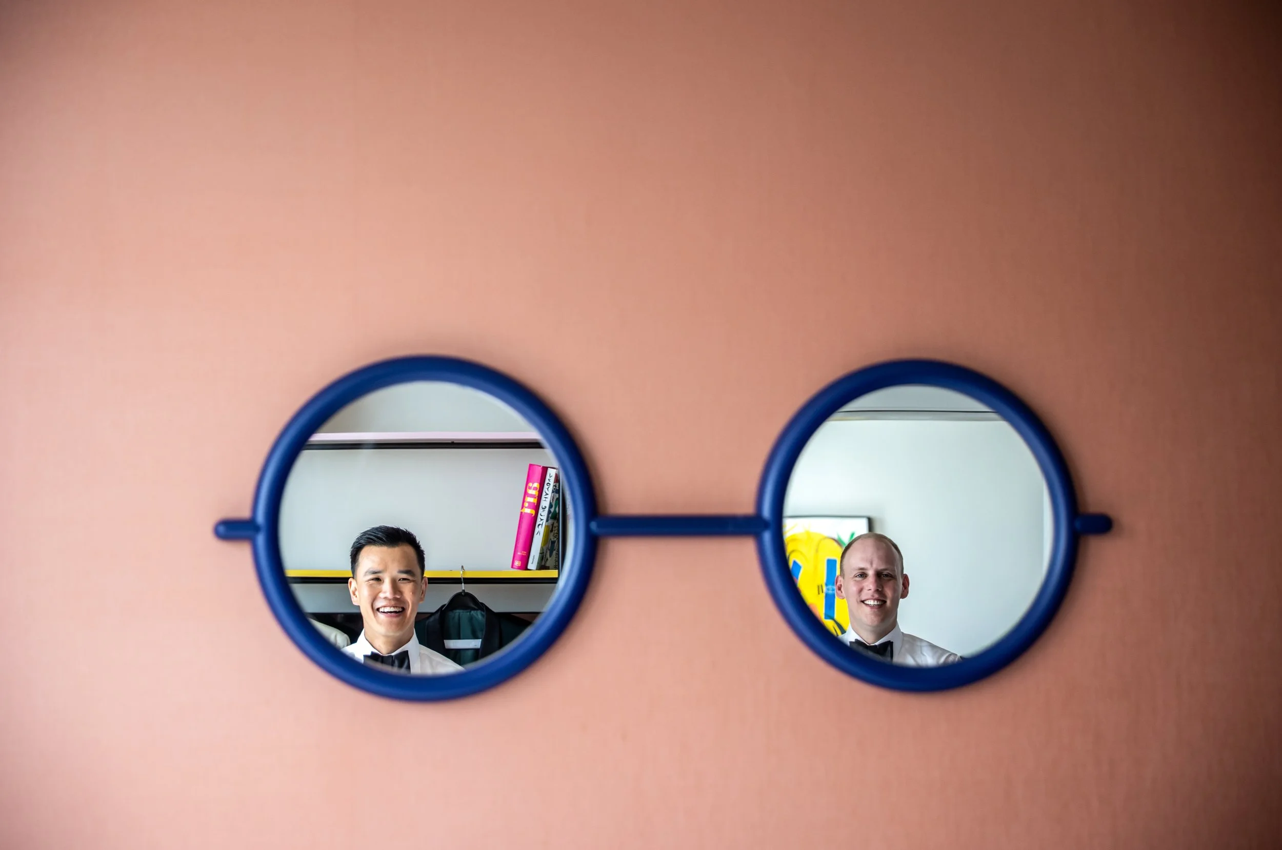 A blue-framed mirror shaped like a pair of glasses on a pink wall. The mirror shows the reflections of two men, one in each "lens" of the glasses. On the left is Kevin, an East Asian man, and on the right is James, a white man, both smiling.