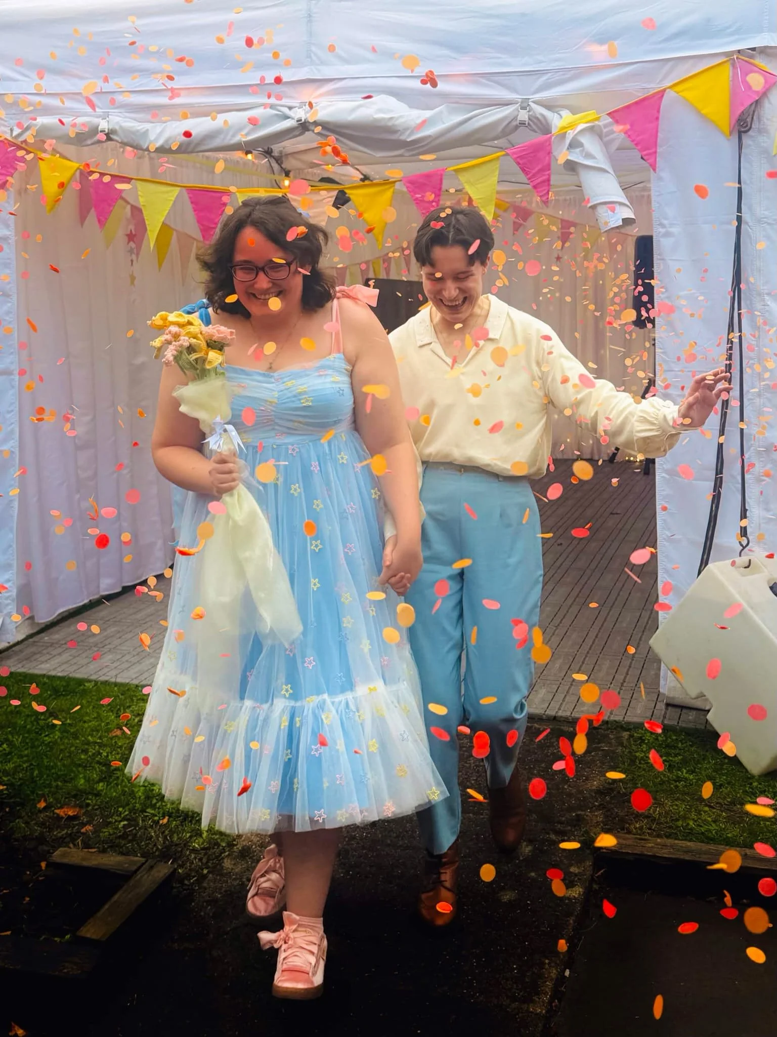 Tabby and Adele walking out of the marquee and being showered in pink and orange confetti. They are both beaming and holding hands.