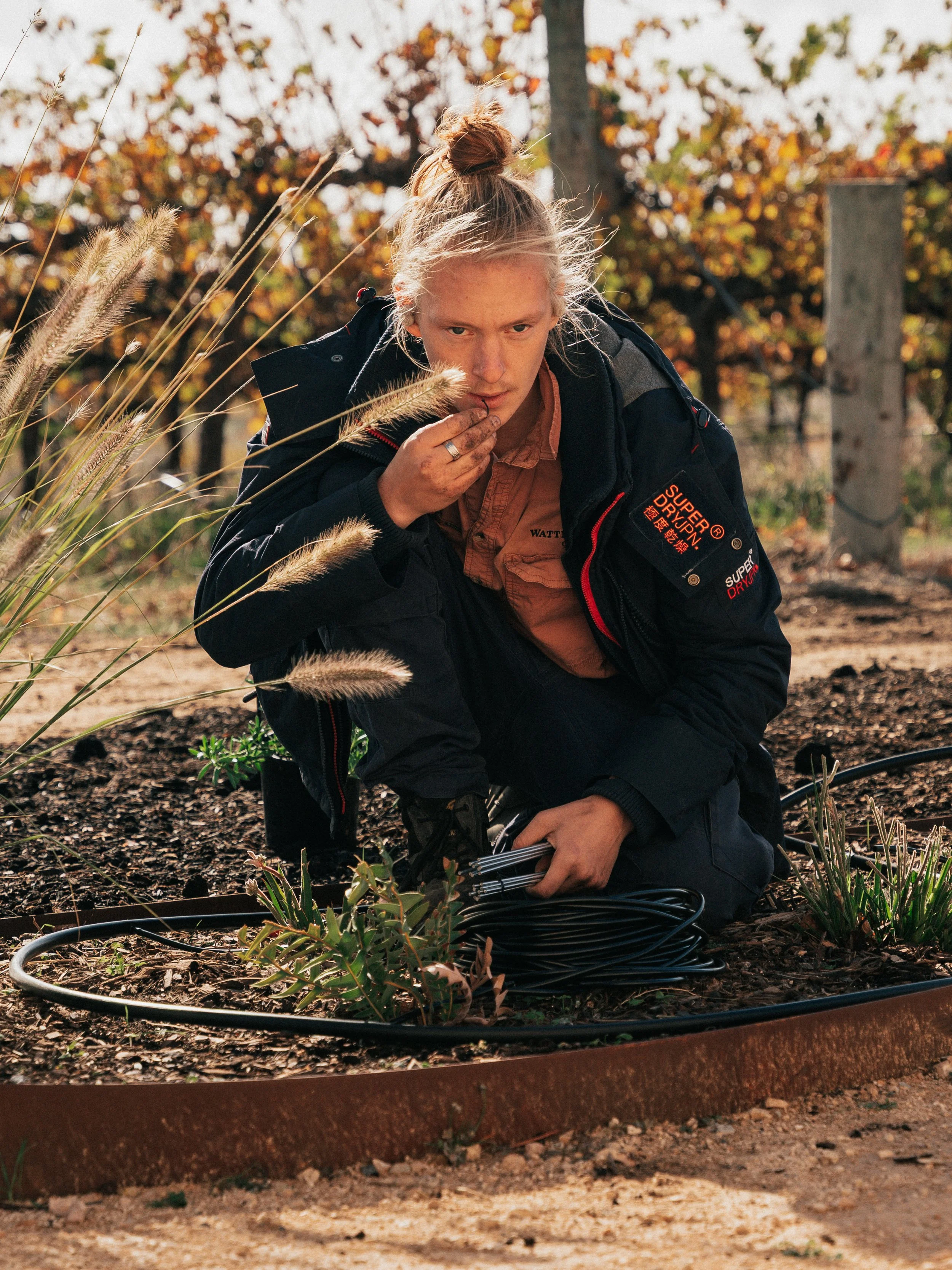 A person with blonde hair tied back kneeling on the ground outdoors, examining plants and holding coiled black cable, surrounded by autumn foliage.