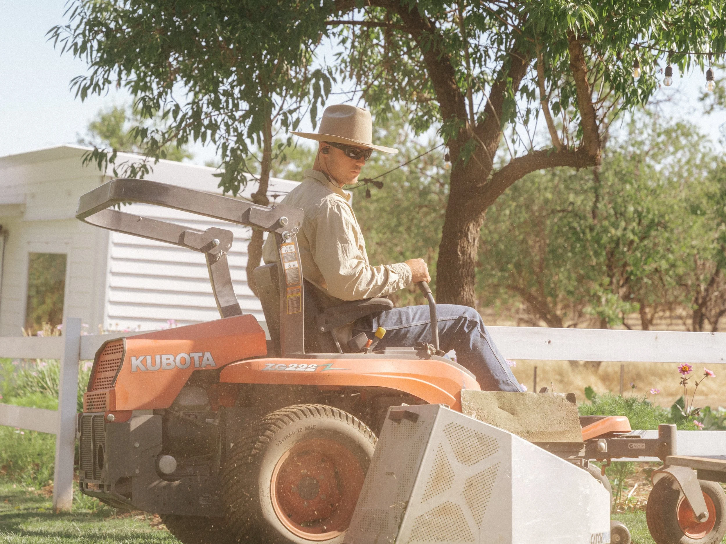 A man wearing a large hat and sunglasses operating a Kubota ZG222 riding lawn mower in a yard with trees and a white house in the background.