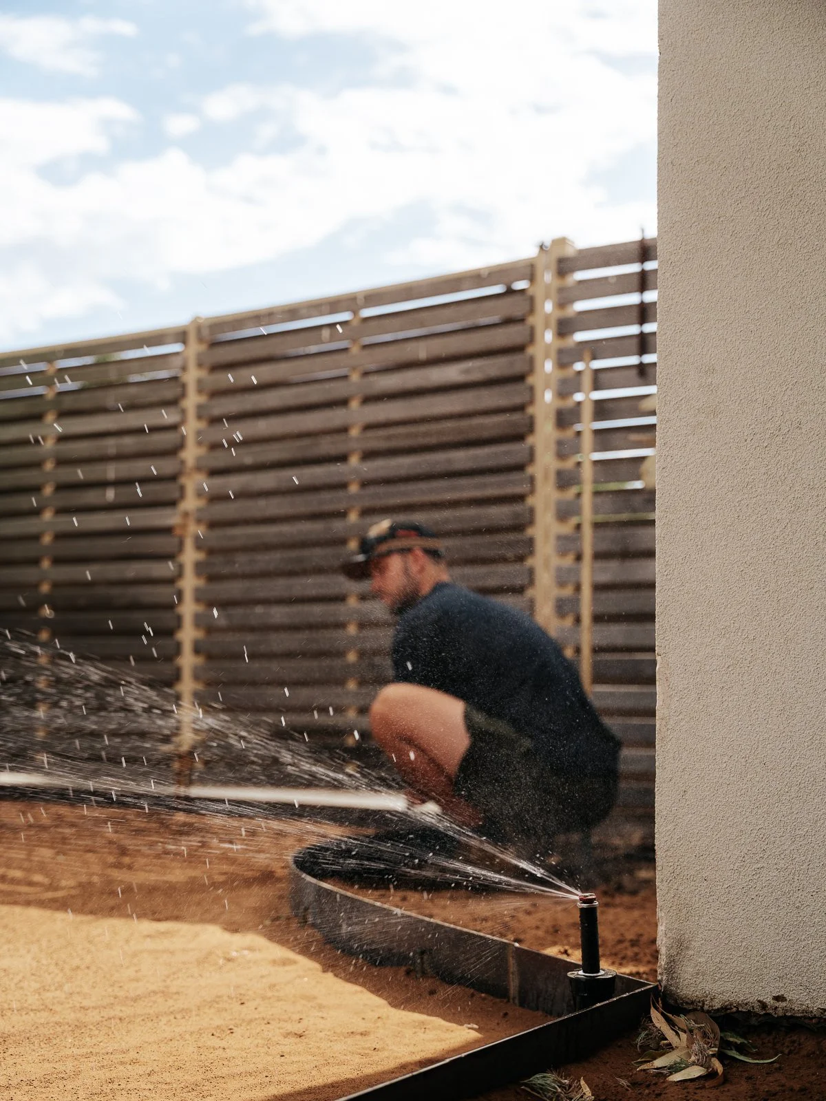 A person crouched near a garden sprinkler on a dirt ground, watering the area beside a white wall with a wooden fence in the background, under a partly cloudy sky.