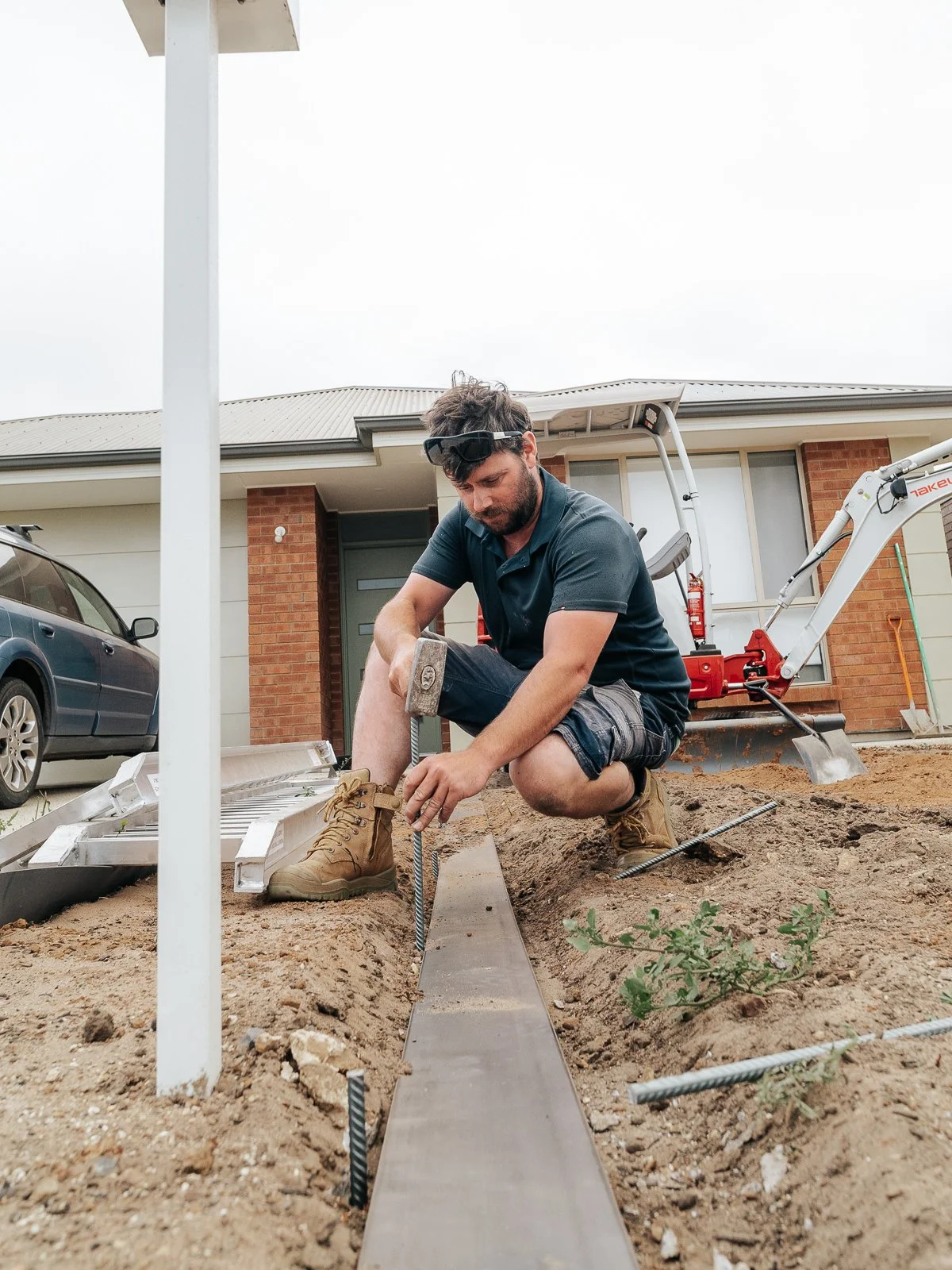 A man working on construction, using a tool on a metal strip in the ground outside a house. He is wearing a black shirt, shorts, boots, and sunglasses resting on his head. Construction equipment is visible in the background.
